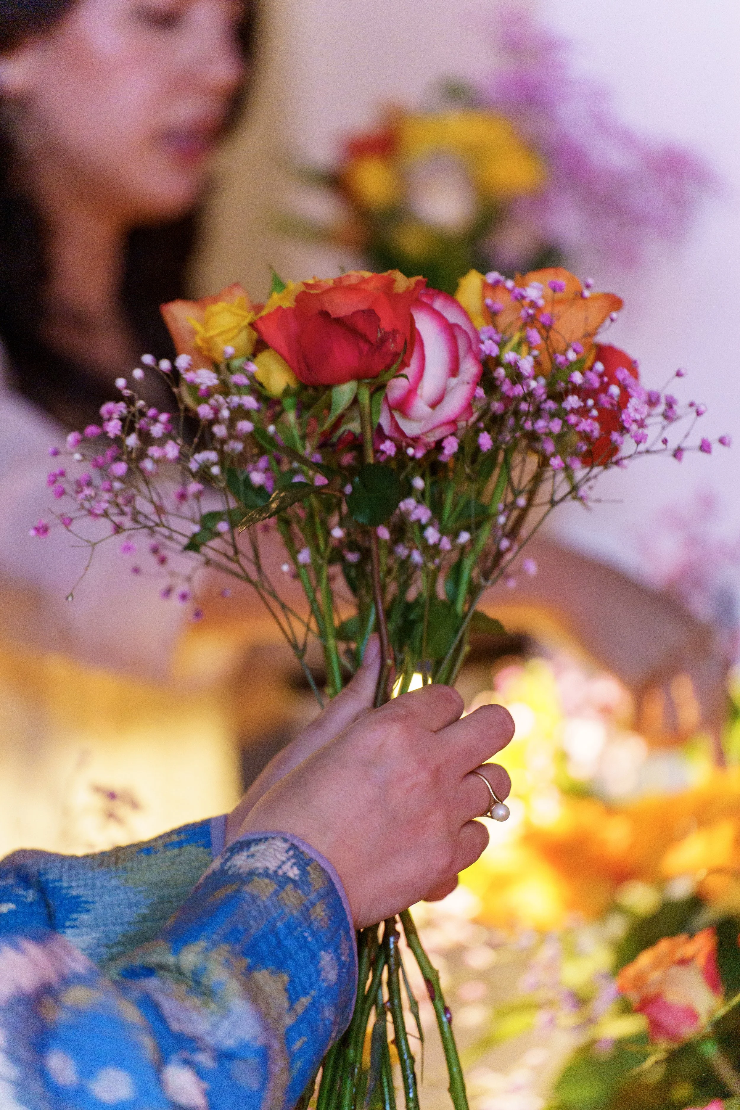 Frau reicht jemandem einen bunten Blumenstrauß mit Rosen, kleinen Blumen und Blättern. Die Person, die den Blumenstrauß hält, trägt einen blauen, gemusterten Ärmel. Der Hintergrund ist unscharf, mit weiteren Blumen und warmem Licht.