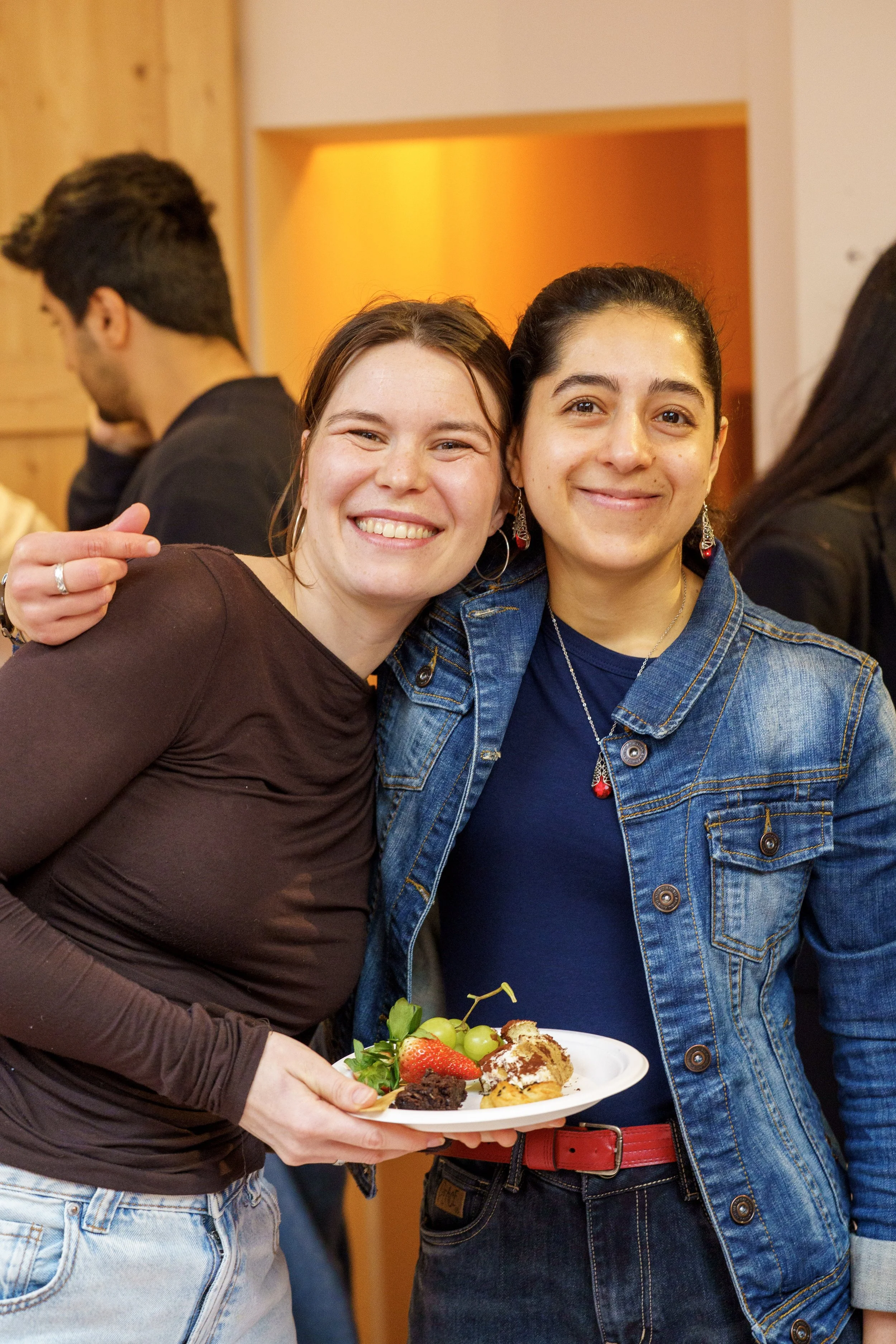 Zwei lachende Frauen posieren bei einer Feier, eine hält einen Teller mit Dessert und Früchten, im Hintergrund sind weitere Menschen.