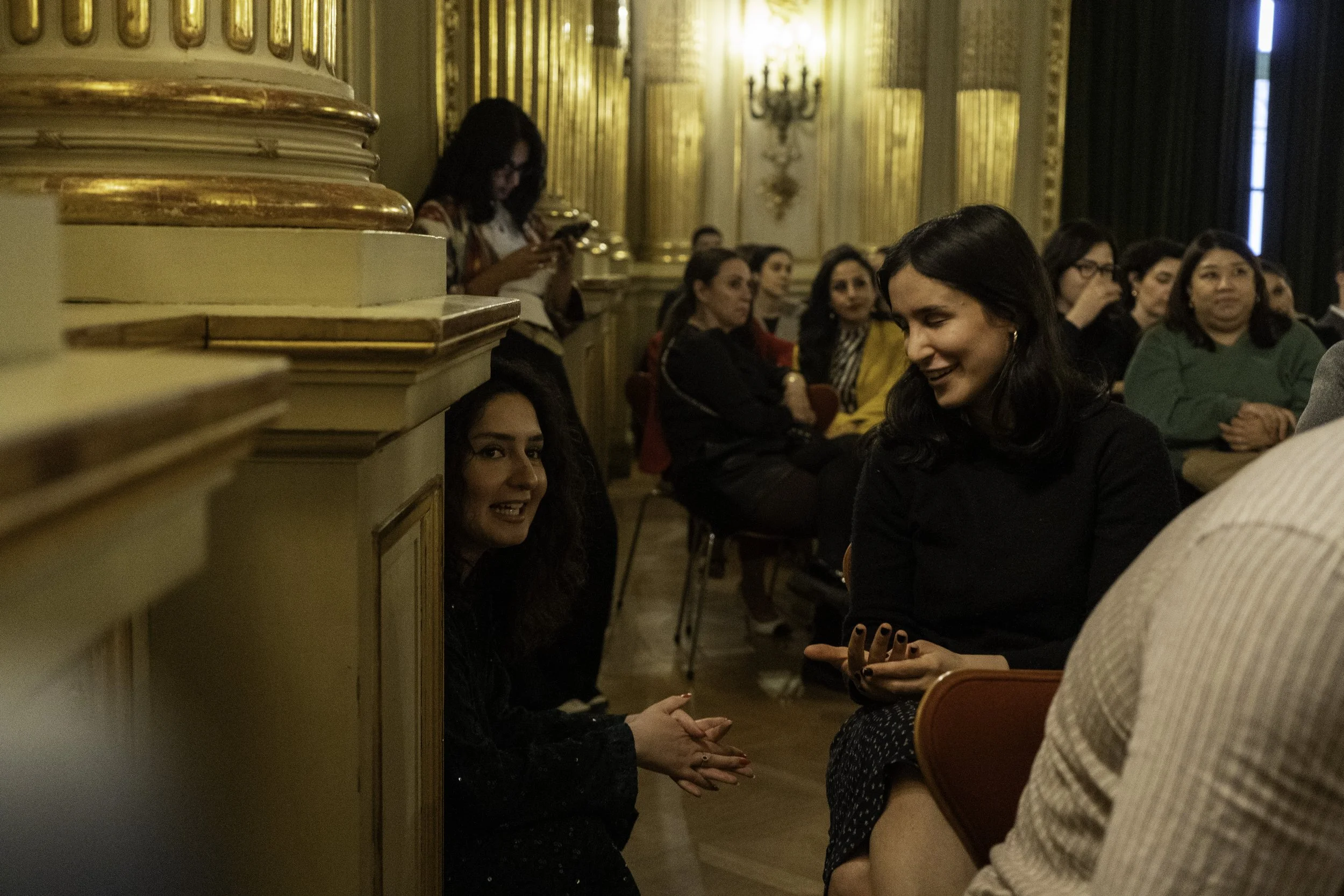 Mehrere Frauen sitzen in einem eleganten Raum mit goldverzierten Wänden, einige lachen und unterhalten sich, während eine Frau im Hintergrund ein Handy benutzt.