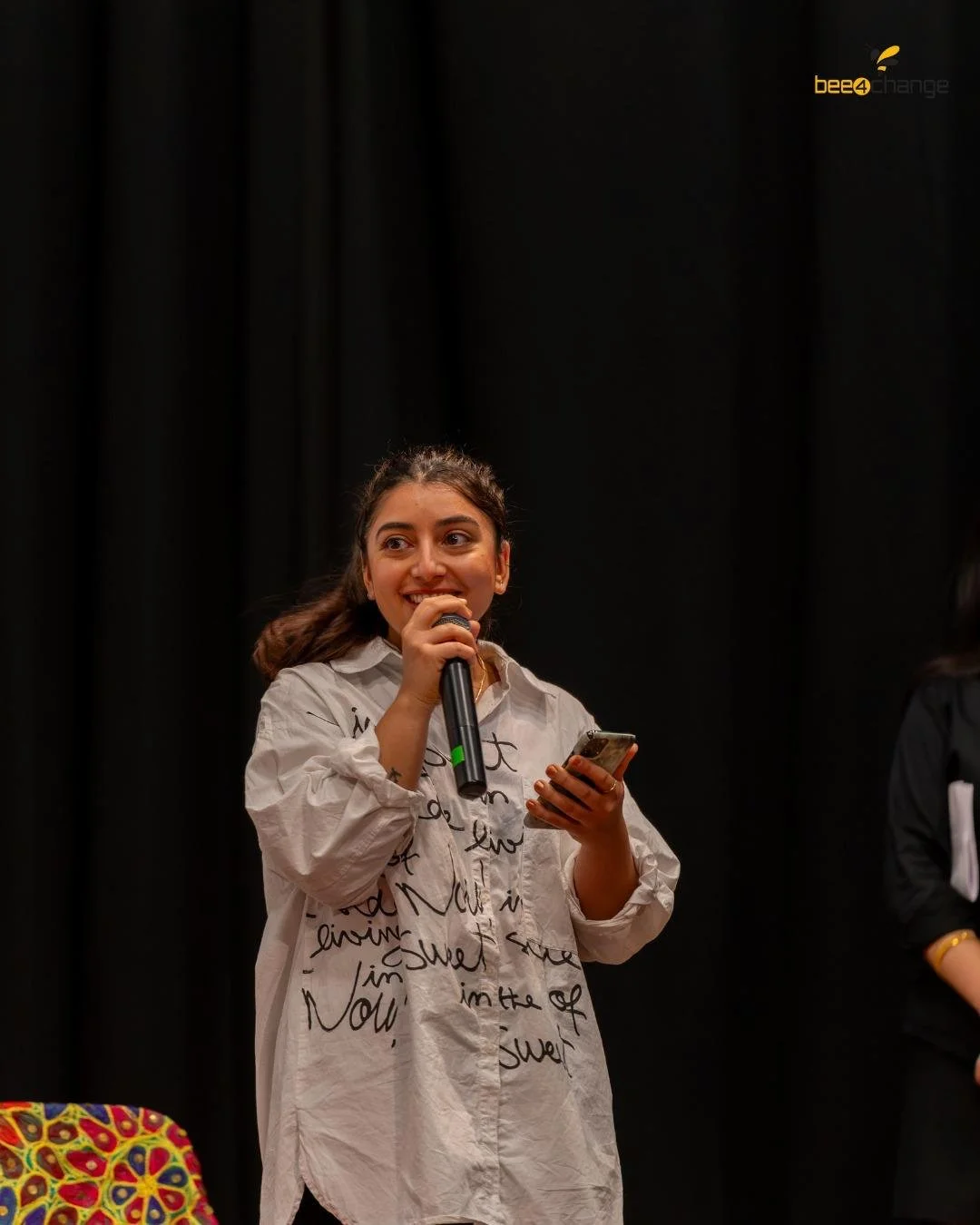 Junge Frau mit Mikrofon und Handy, spricht auf einer Bühne mit schwarzem Hintergrund.