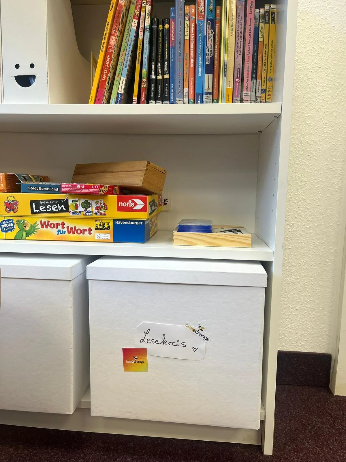 Ein Bücherregal mit bunten Kinderbüchern, einer kleinen Holzkiste und Spielmaterialien. Darunter ein weißer Karton mit einem Aufkleber, auf dem "Lesekreis" geschrieben steht.