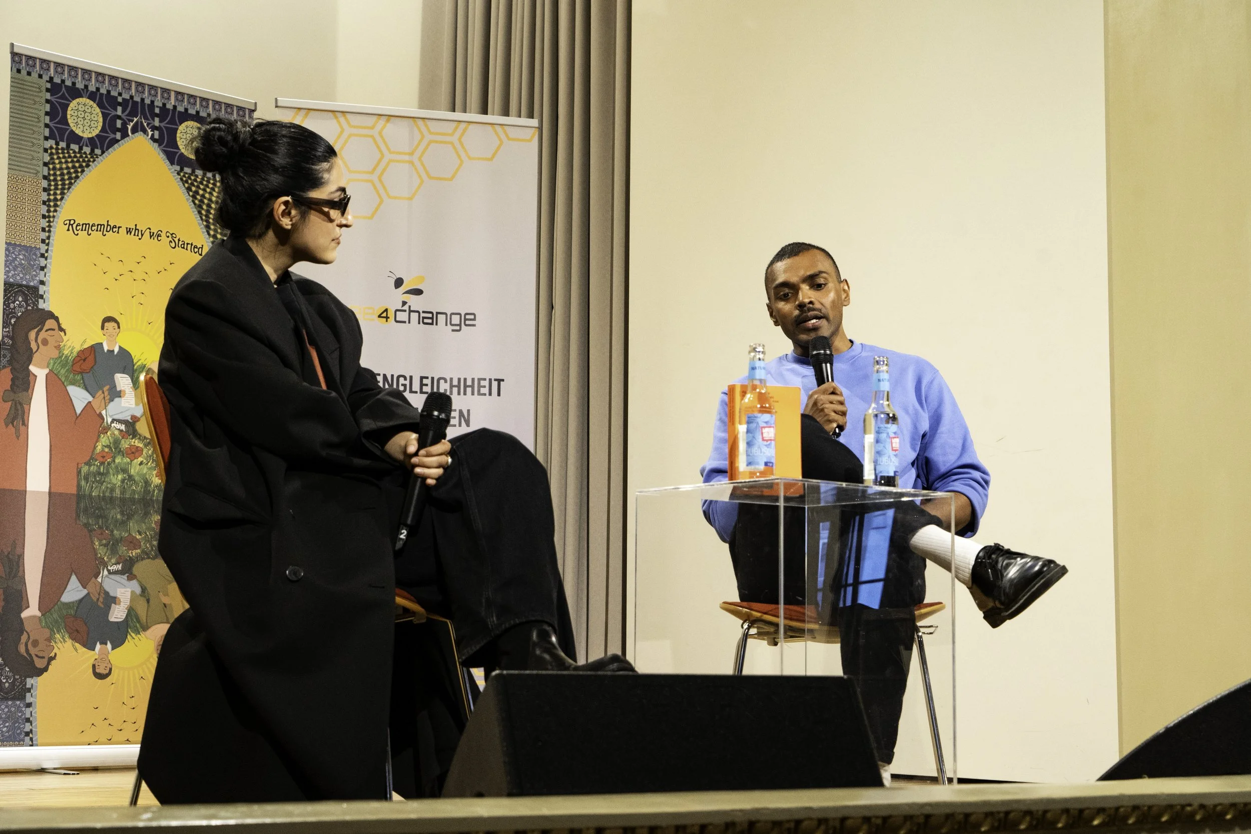 Zwei Personen bei einer Diskussion auf einer Bühne, eine Frau sitzt mit Mikrofon, ein Mann sitzt mit Mikrofon und hat einen Beinstuhl, im Hintergrund Banner mit Text und Illustrationen.