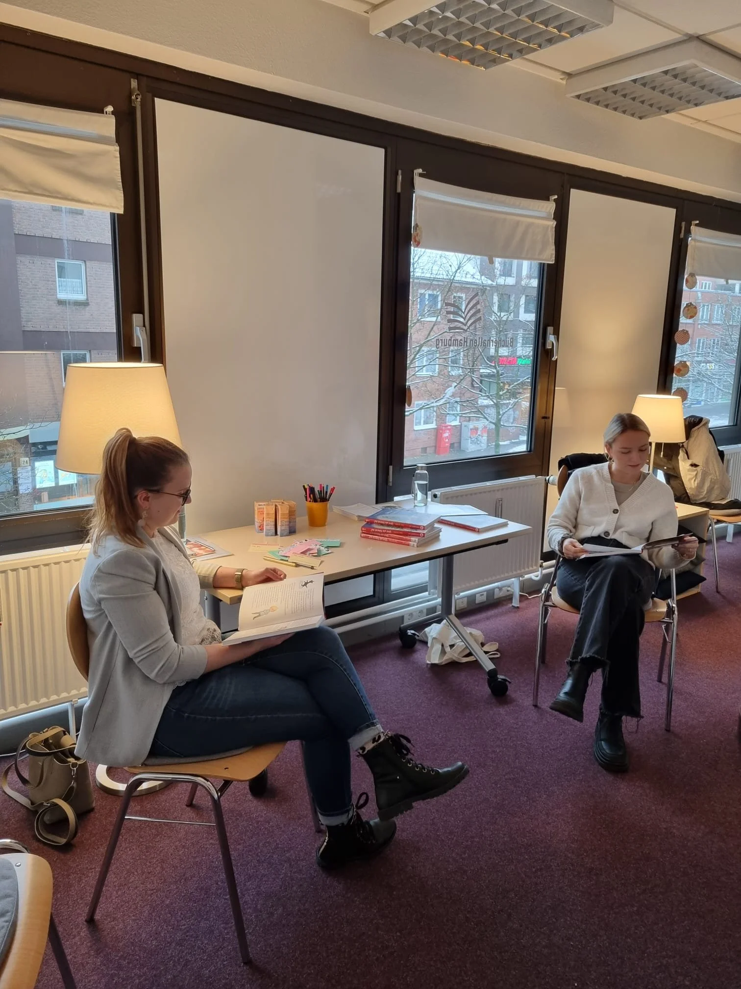 Zwei Frauen sitzen in einem Raum mit großen Fenstern, lesen Bücher und unterhalten sich. Es gibt einen Tisch mit Büchern, Schreibutensilien und Papier, sowie Decken und Lampen im Hintergrund.