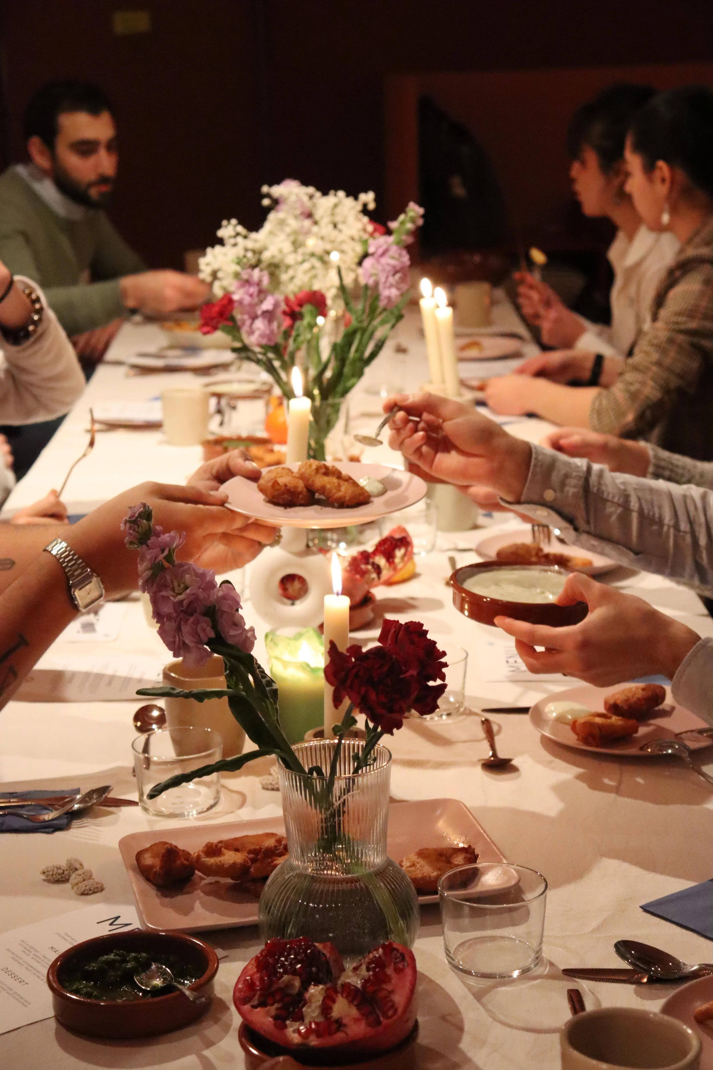 Menschen sitzen an einem festlichen Abendessen um einen Tisch mit Kerzen, Blumen und verschiedenen Speisen.