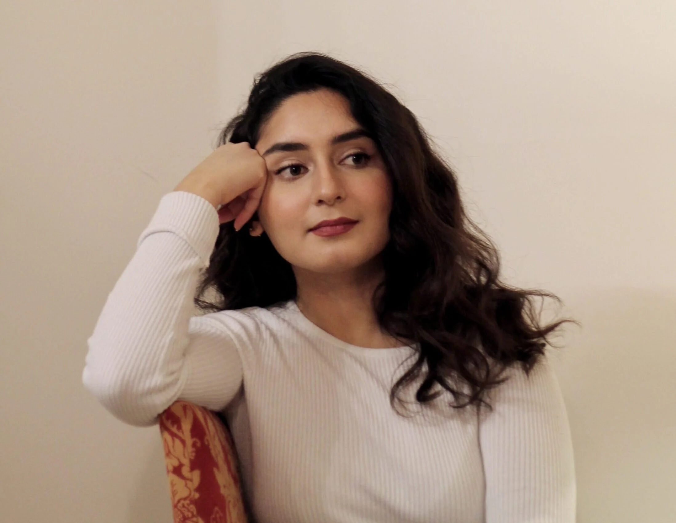 Eine junge Frau mit dunklen, lockigen Haaren, die auf der linken Seite ihres Gesichts liegt, sitzt an einer roten, bestickten Stuhllehne, trägt ein weißes langärmliges Oberteil und schaut nach rechts, mit einer neutralen Gesichtsausdruck vor einem cremefarbenen Hintergrund.