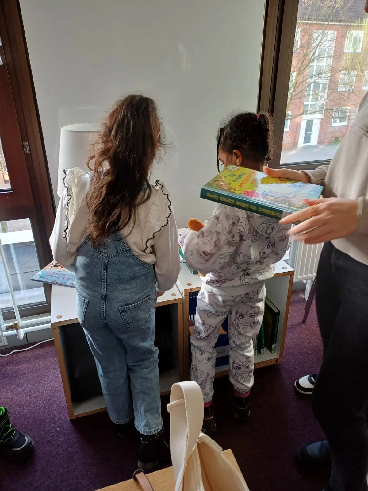 Zwei Kinder stehen vor einem Bücherregal und schauen sich Bücher an, während eine erwachsene Person ein Buch hält.
