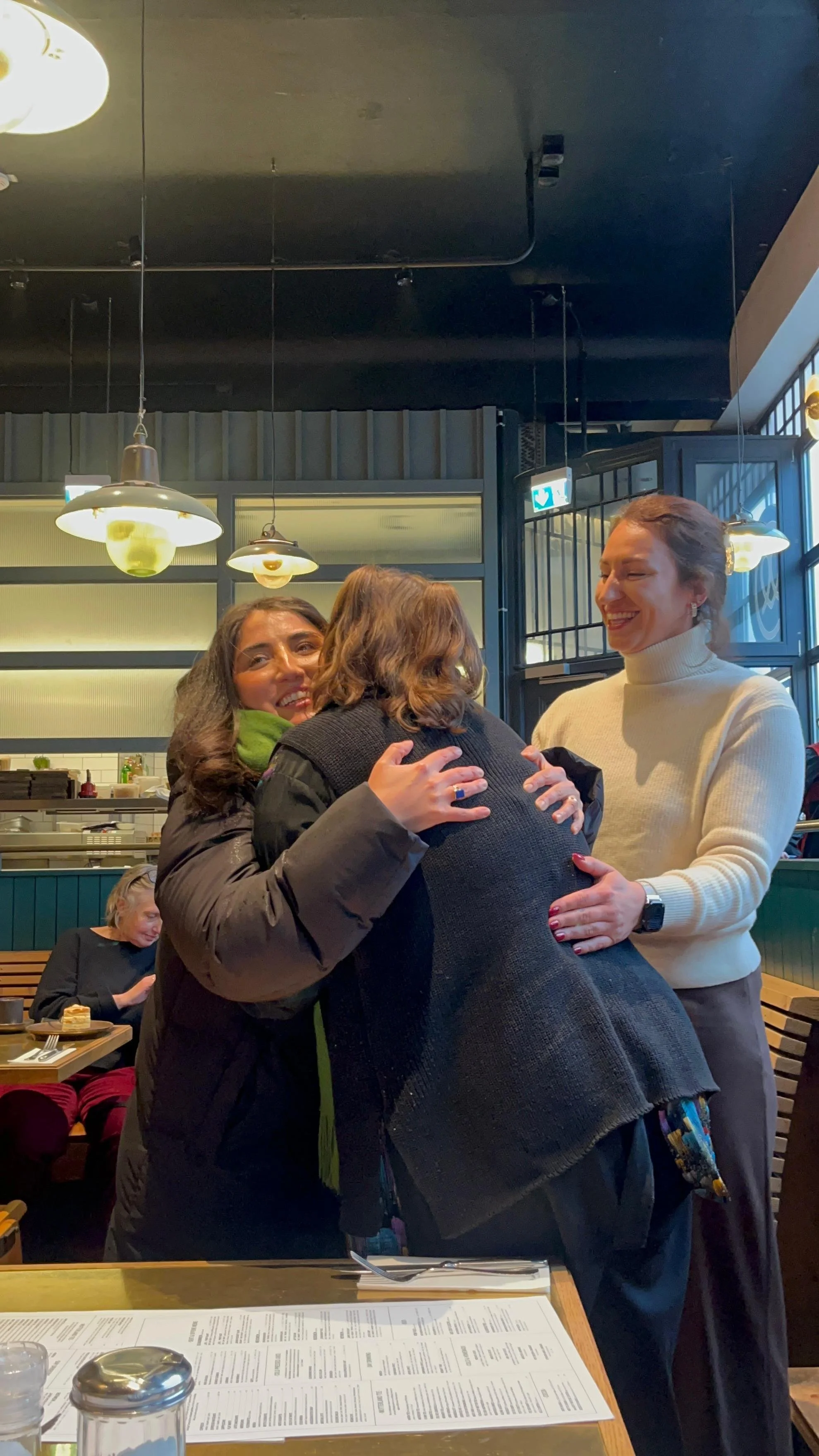Drei Frauen umarmen sich in einem Restaurant, lachen, im Hintergrund eine andere Frau, die essen hat.