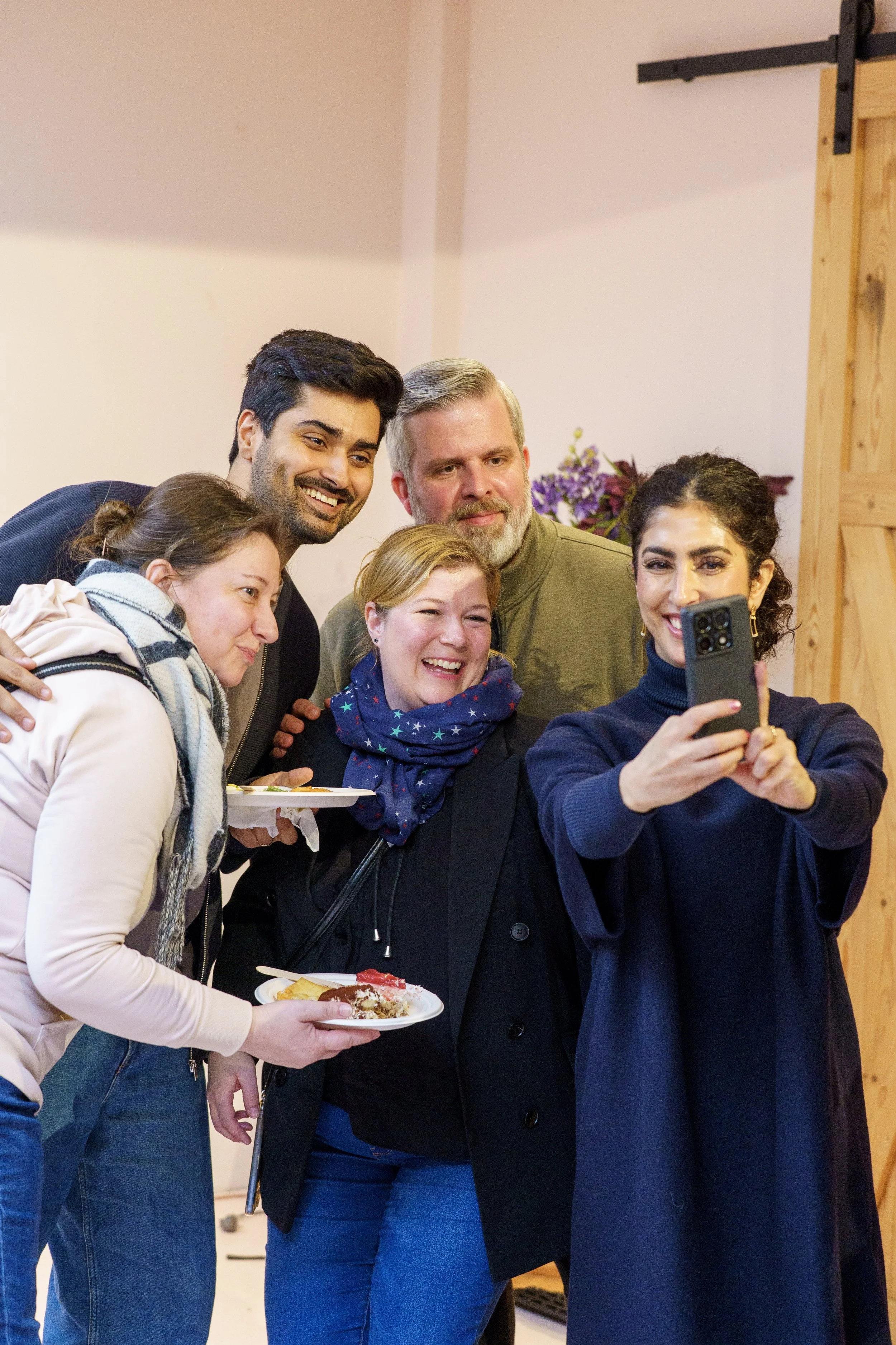 Menschen machen ein Selfie bei einer Feier, einer hält ein Smartphone, zwei halten Teller mit Essen, sie lachen und posieren gemeinsam.