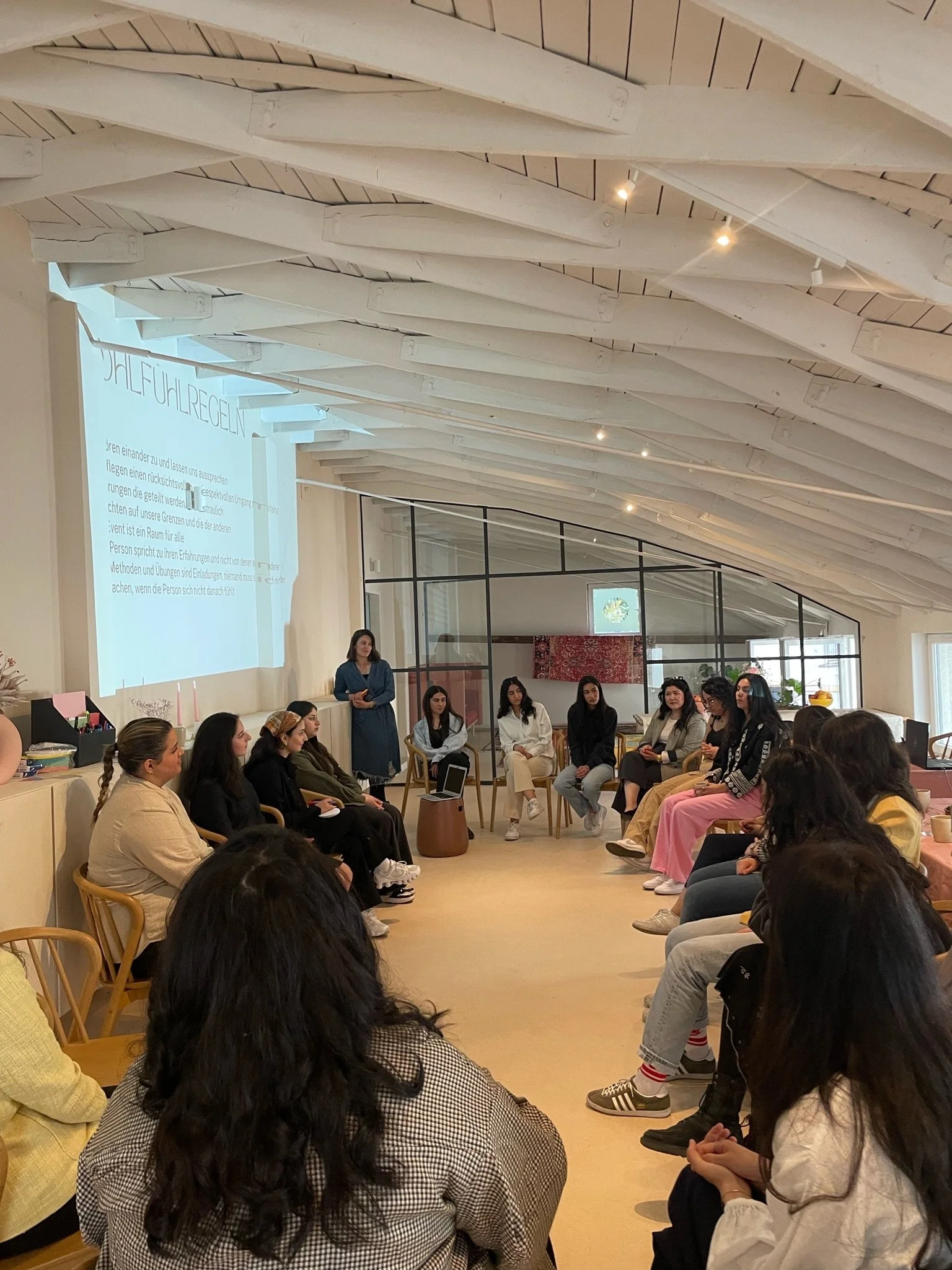 Eine Gruppe von Menschen sitzt in einem Kreis in einem hellen Raum und hört einer Frau zu, die vorne steht. Es sieht nach einem Workshop oder Seminar aus.