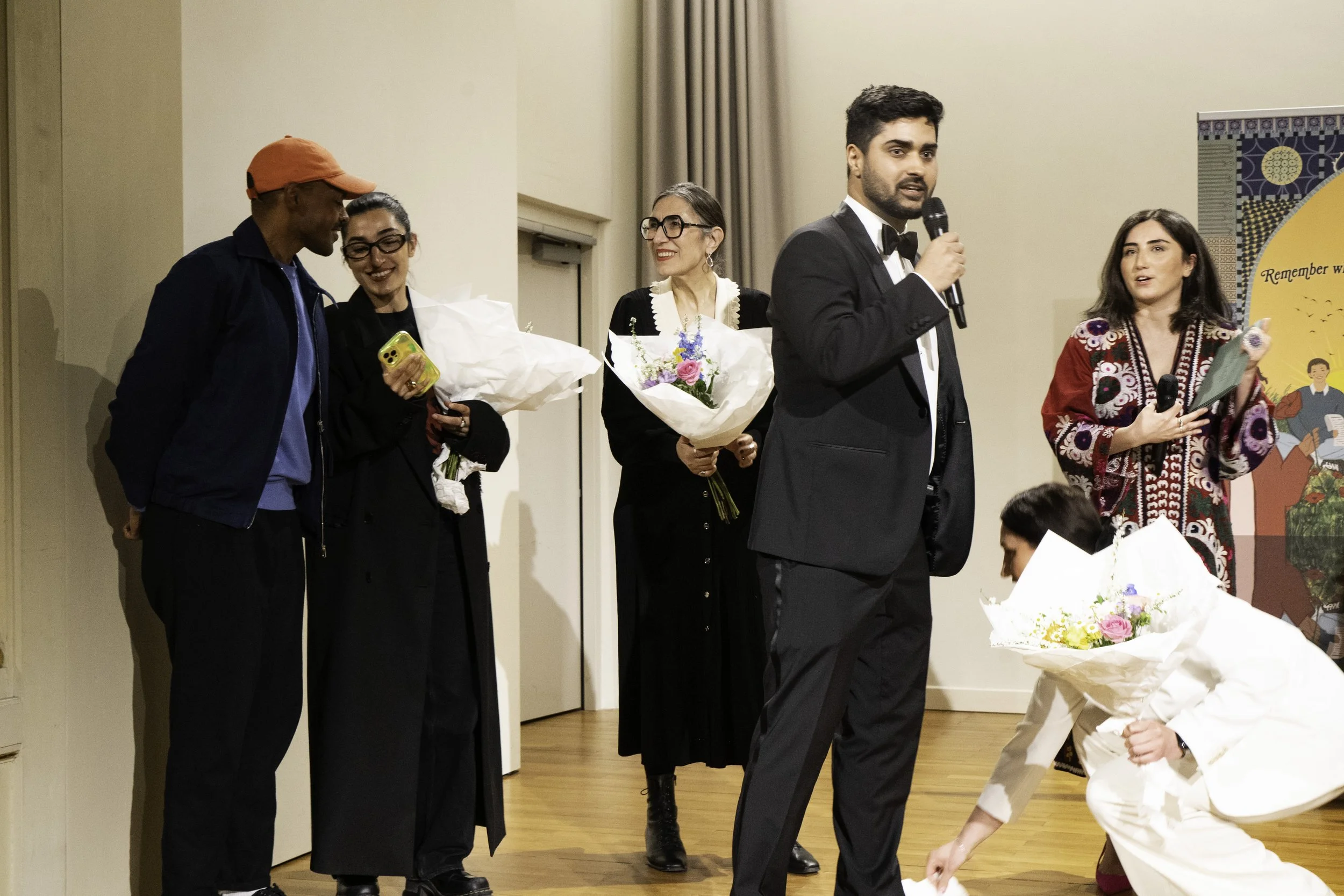 Menschen auf einer Bühne bei einer Veranstaltung, vier Frauen mit Blumenstrauß und einem Mann in einem schwarzen Anzug, alle halten Mikrofone und sprechen.