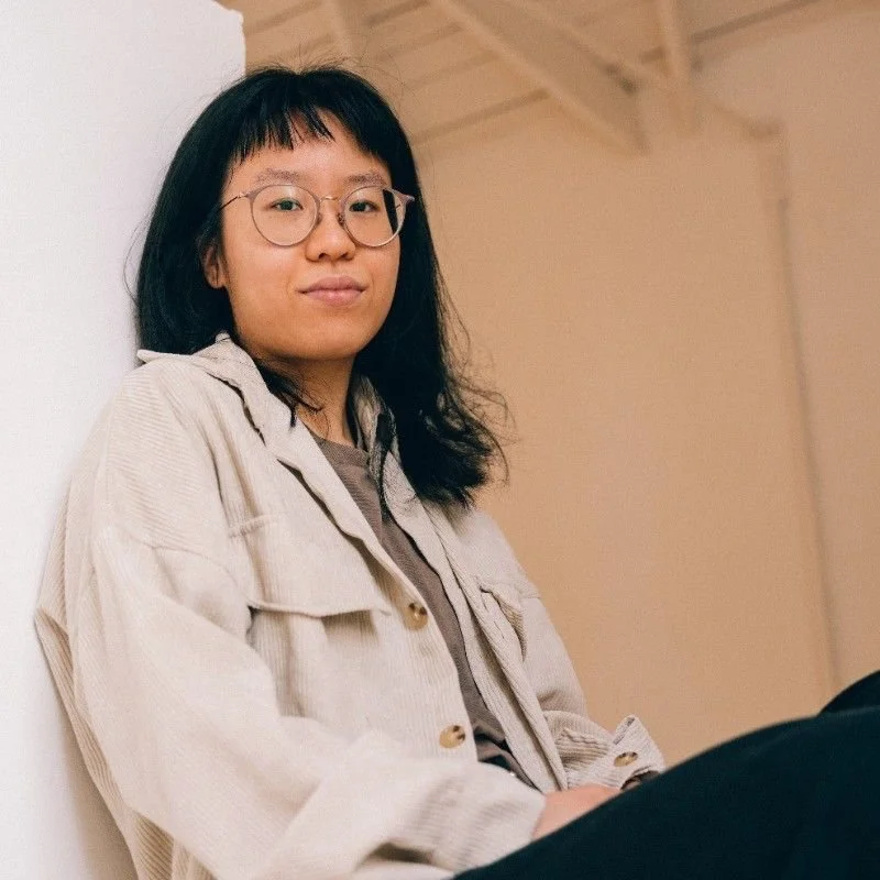 Junge Frau mit Brille lehnt an einer weißen Wand, lächelnd in einem Raum mit Holzdecke.