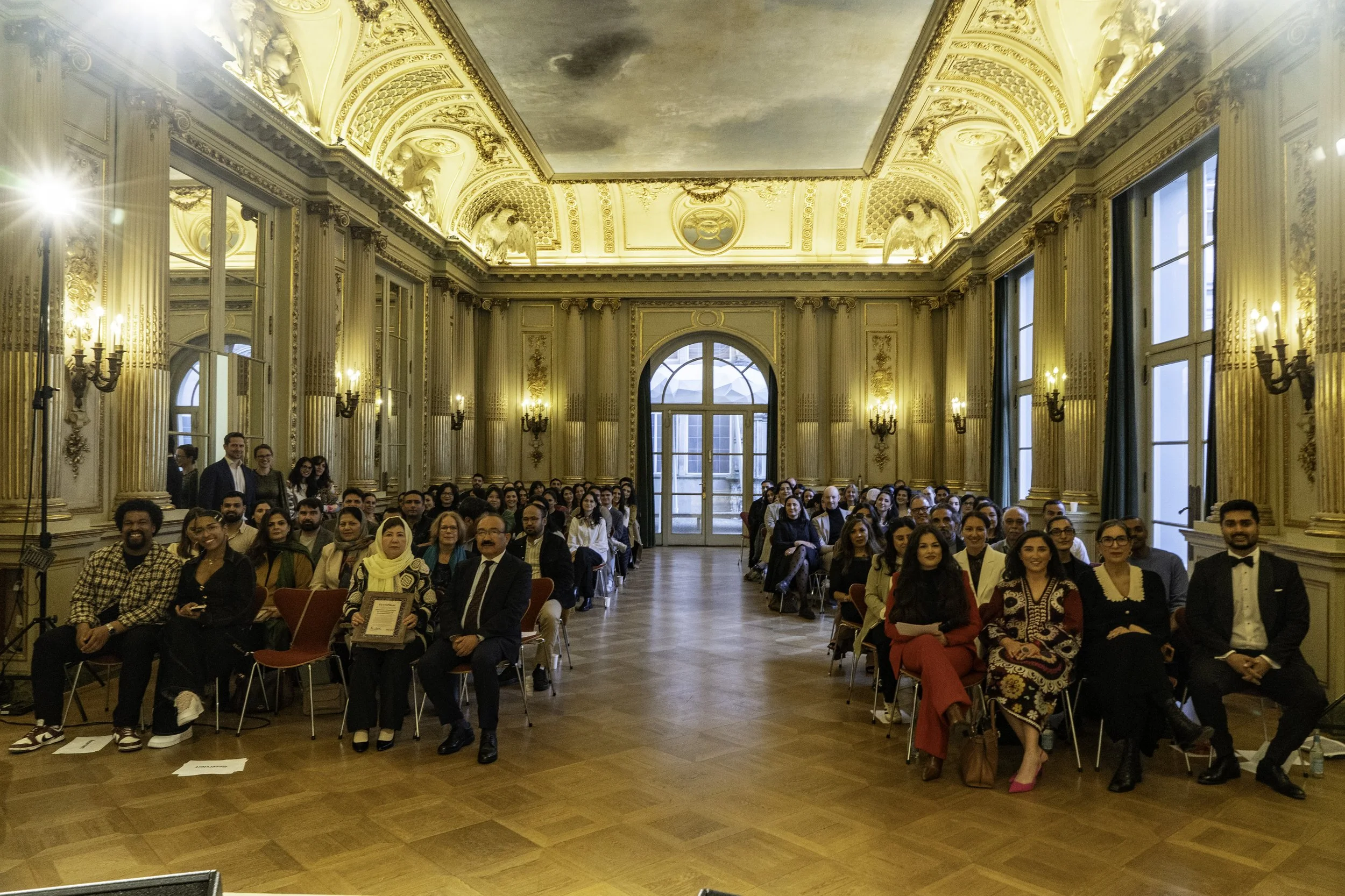 Eine große Gruppe von Menschen sitzt in einem eleganten, goldverzierten Saal mit hohen Fenstern und ornamentalen Deckenledern.