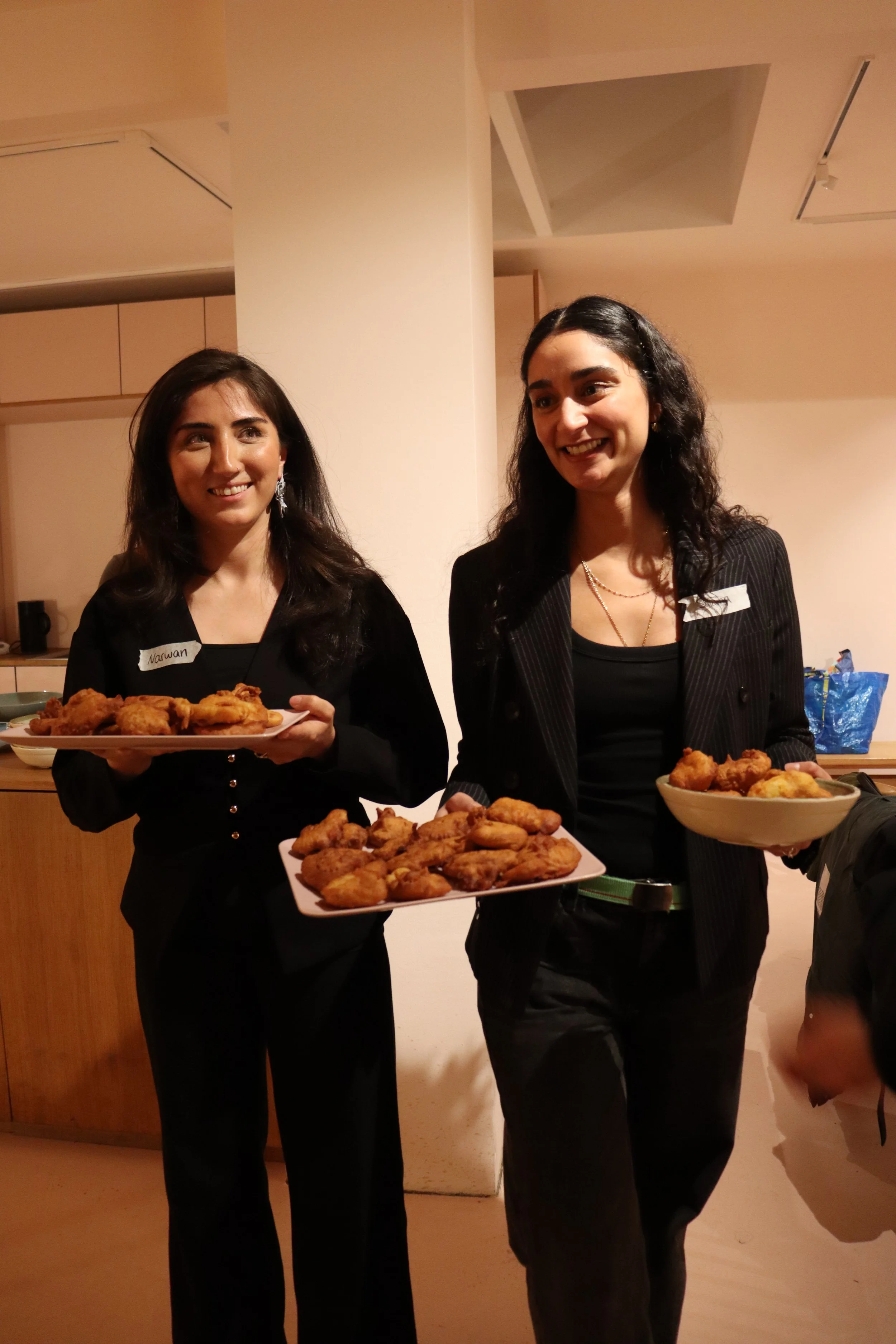 Zwei Frauen in schwarzer Kleidung halten Teller mit frittiertem Essen bei einer Veranstaltung.