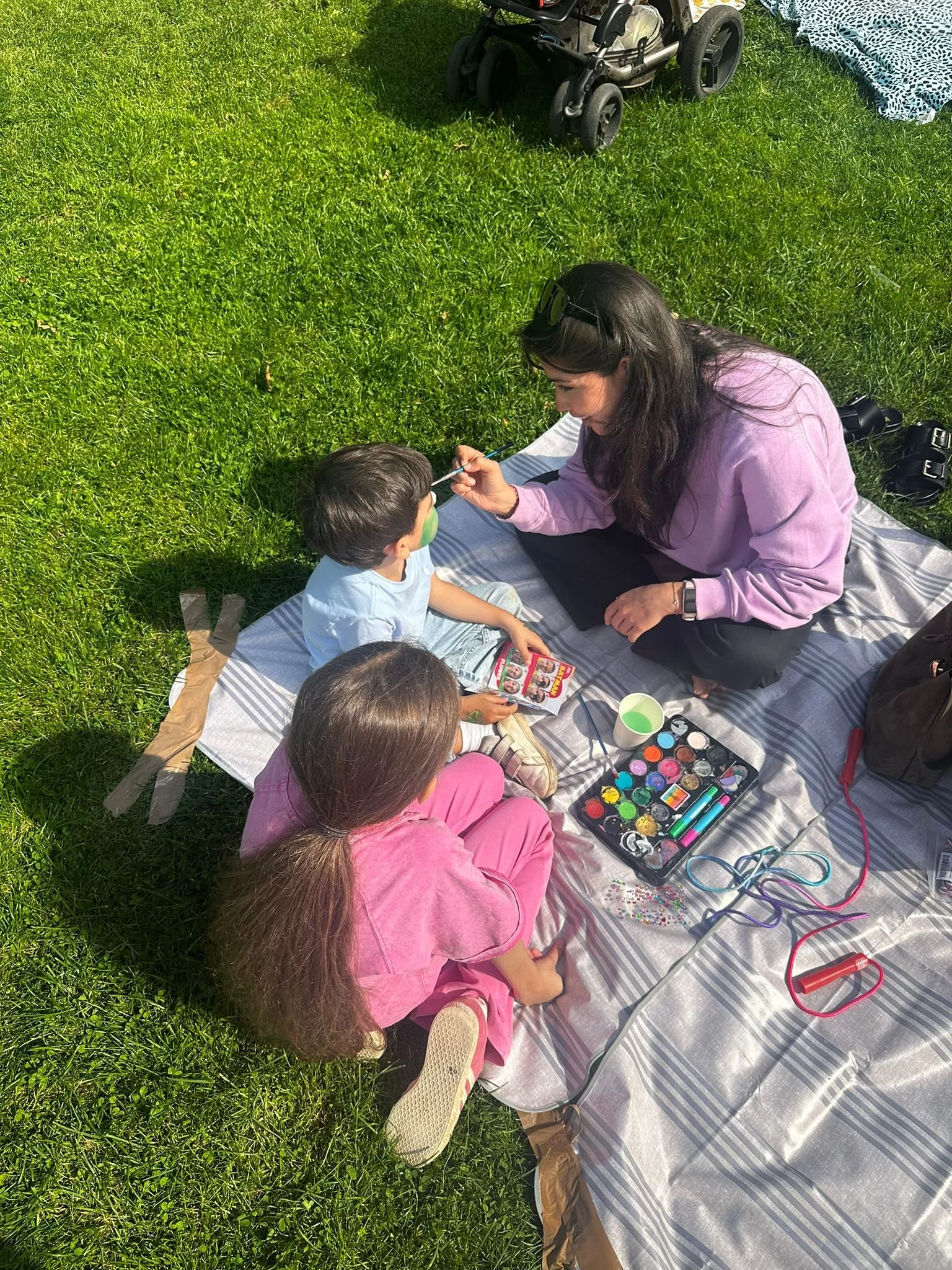 Mutter malt mit Pinsel Gesicht eines Kindes beim Picknick im Park, Kinder sitzen auf einer Decke mit Make-up-Set, Umgebung ist grün mit Gras und Kinderwagen im Hintergrund.