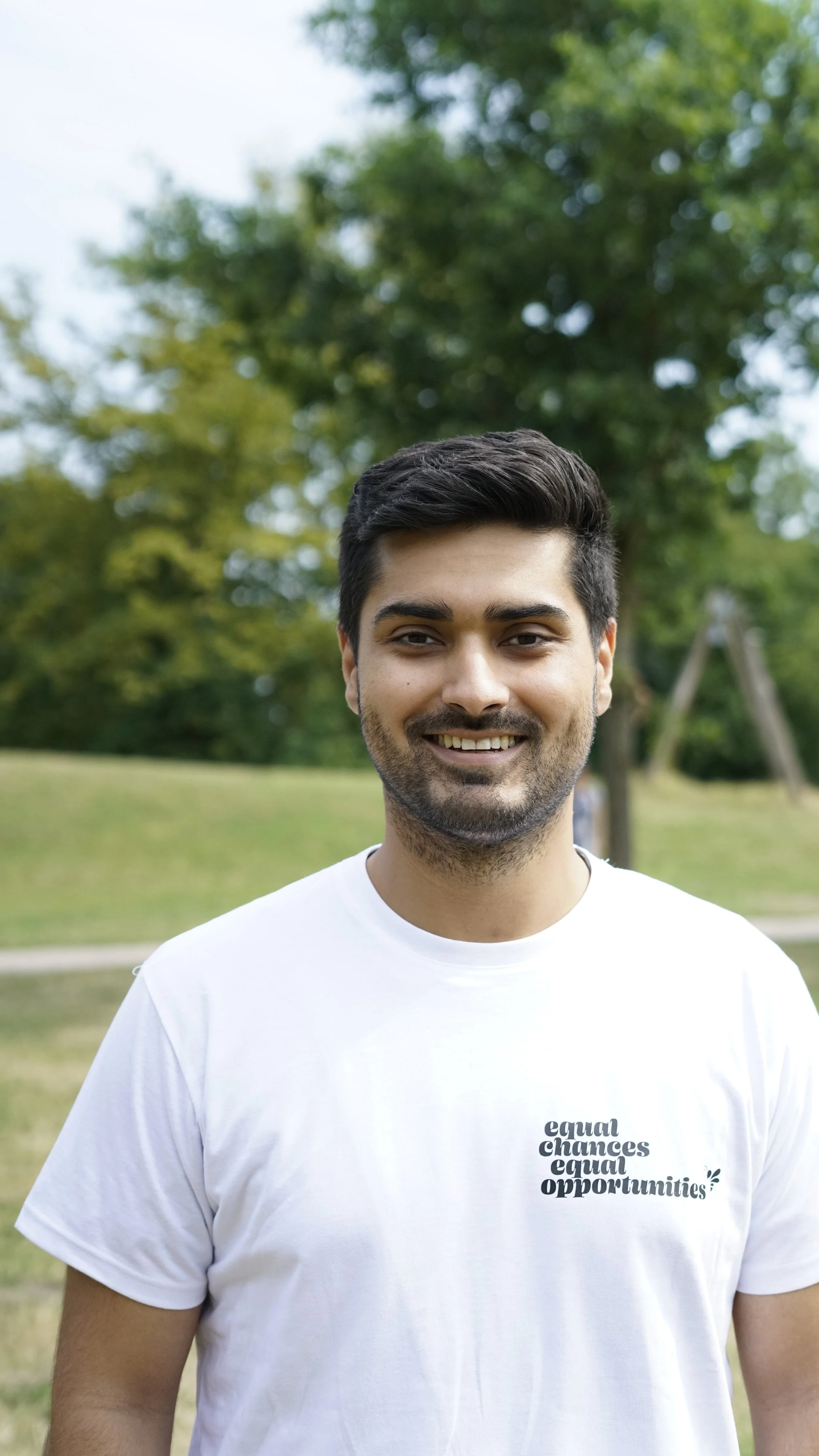 Ein lachender junger Mann steht im Park, trägt ein weißes T-Shirt mit dem Text 'equal chances equal opportunities', im Hintergrund Bäume und eine Schaukel.
