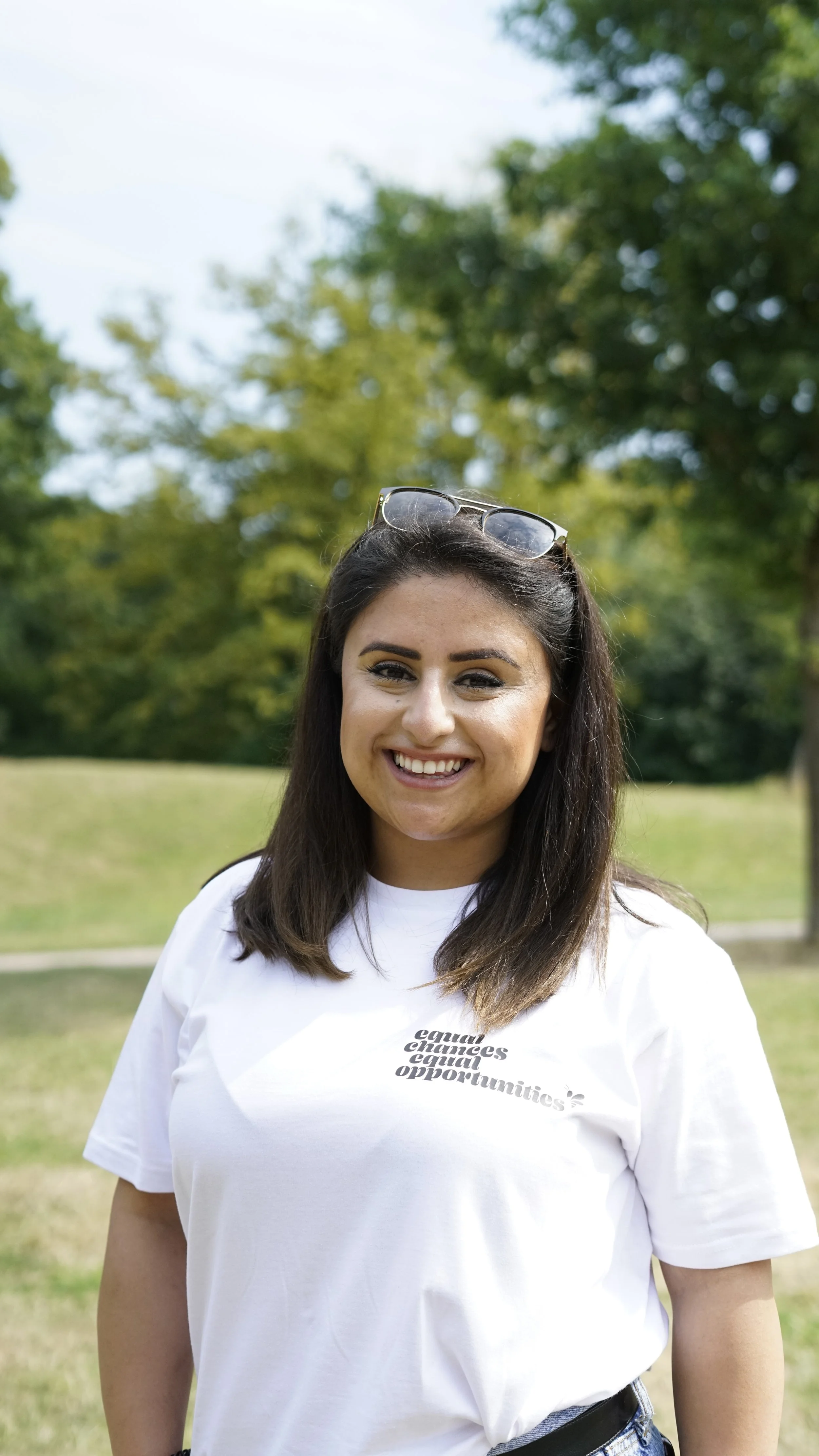 Junge Frau mit dunklem Haar trägt weiße T-Shirt mit Text und Sonnenbrille auf dem Kopf, lächelnd im Park im Freien mit Bäumen im Hintergrund.
