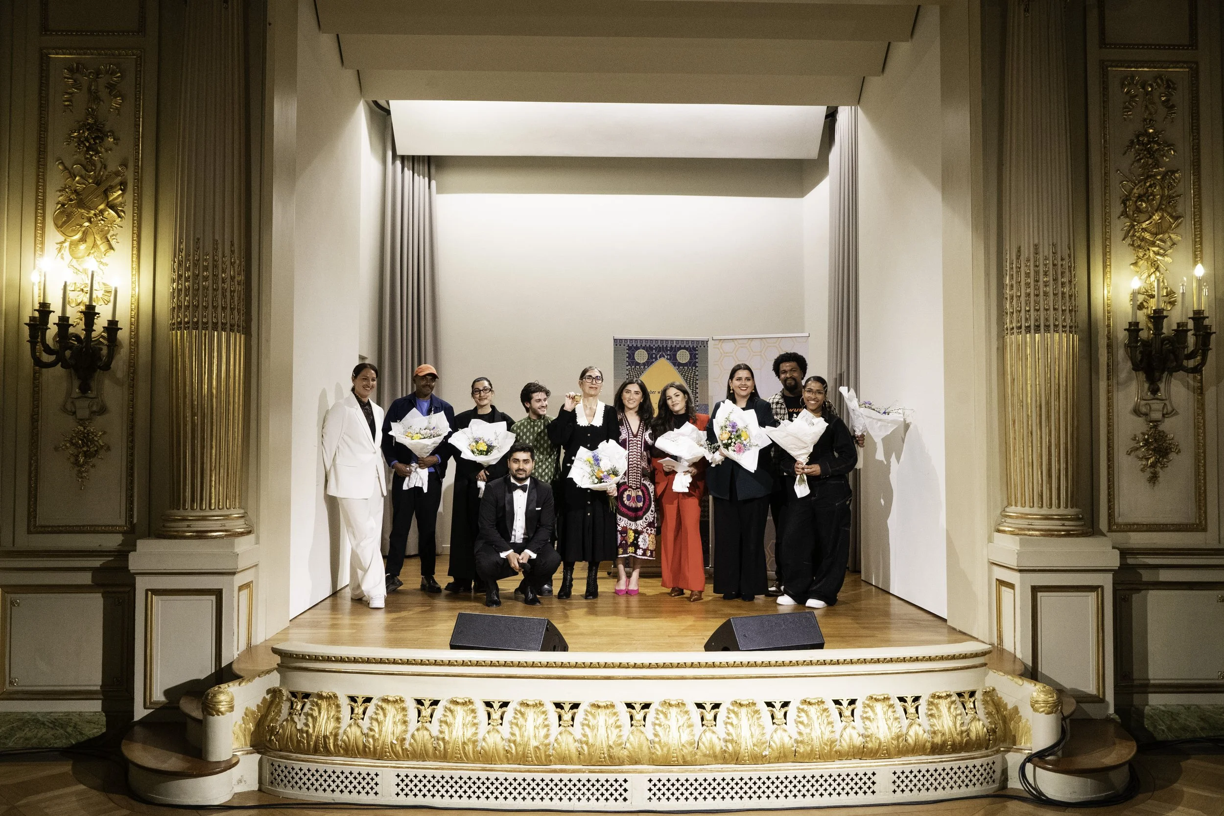 Gruppe von Menschen auf einer Bühne in einem eleganten Saal, einige halten Blumen, alle lächeln. Es sieht aus wie eine Veranstaltung oder Feier.