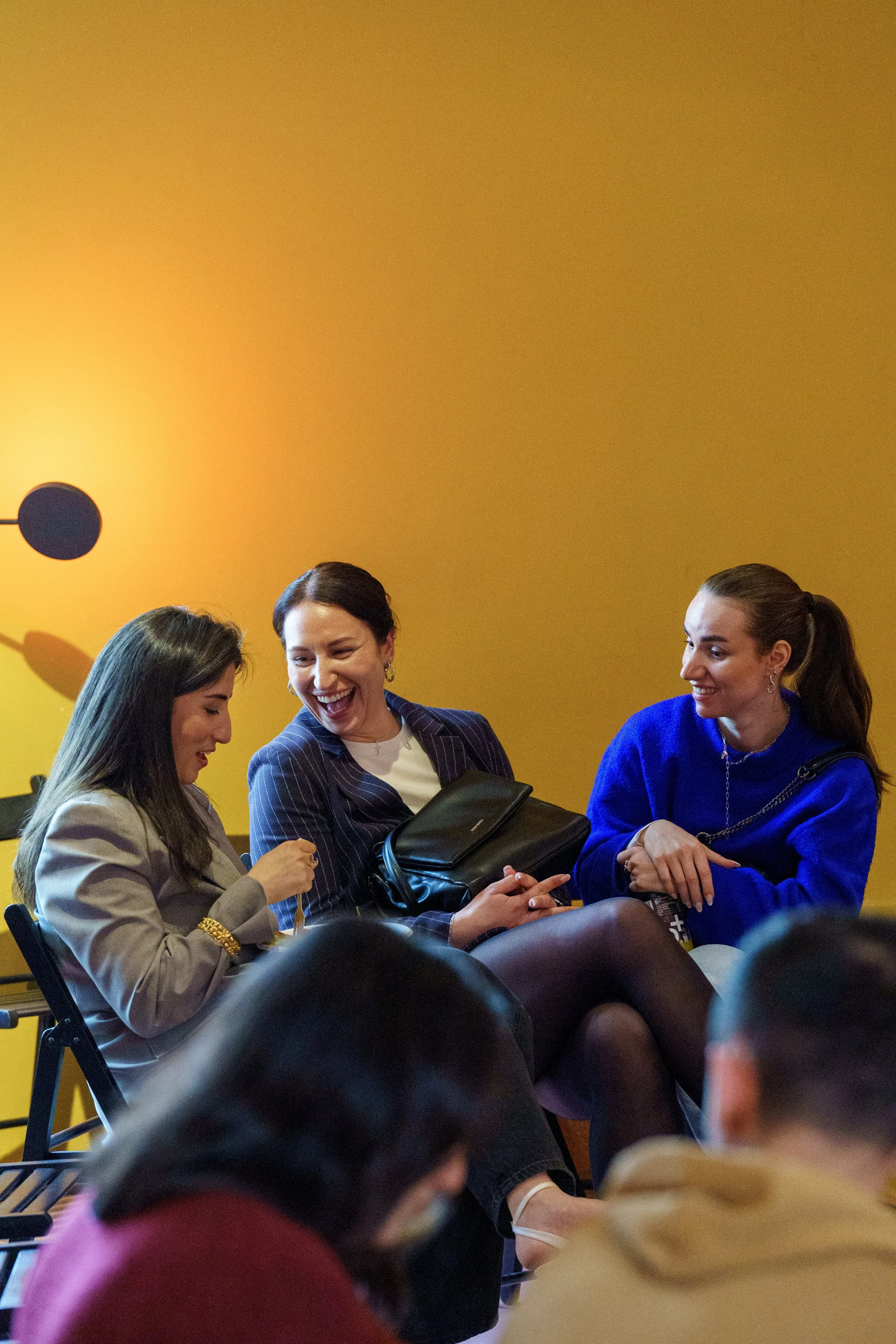 Drei Frauen sitzen zusammen, lachen und sprechen in einem Raum mit gelber Wand im Hintergrund.