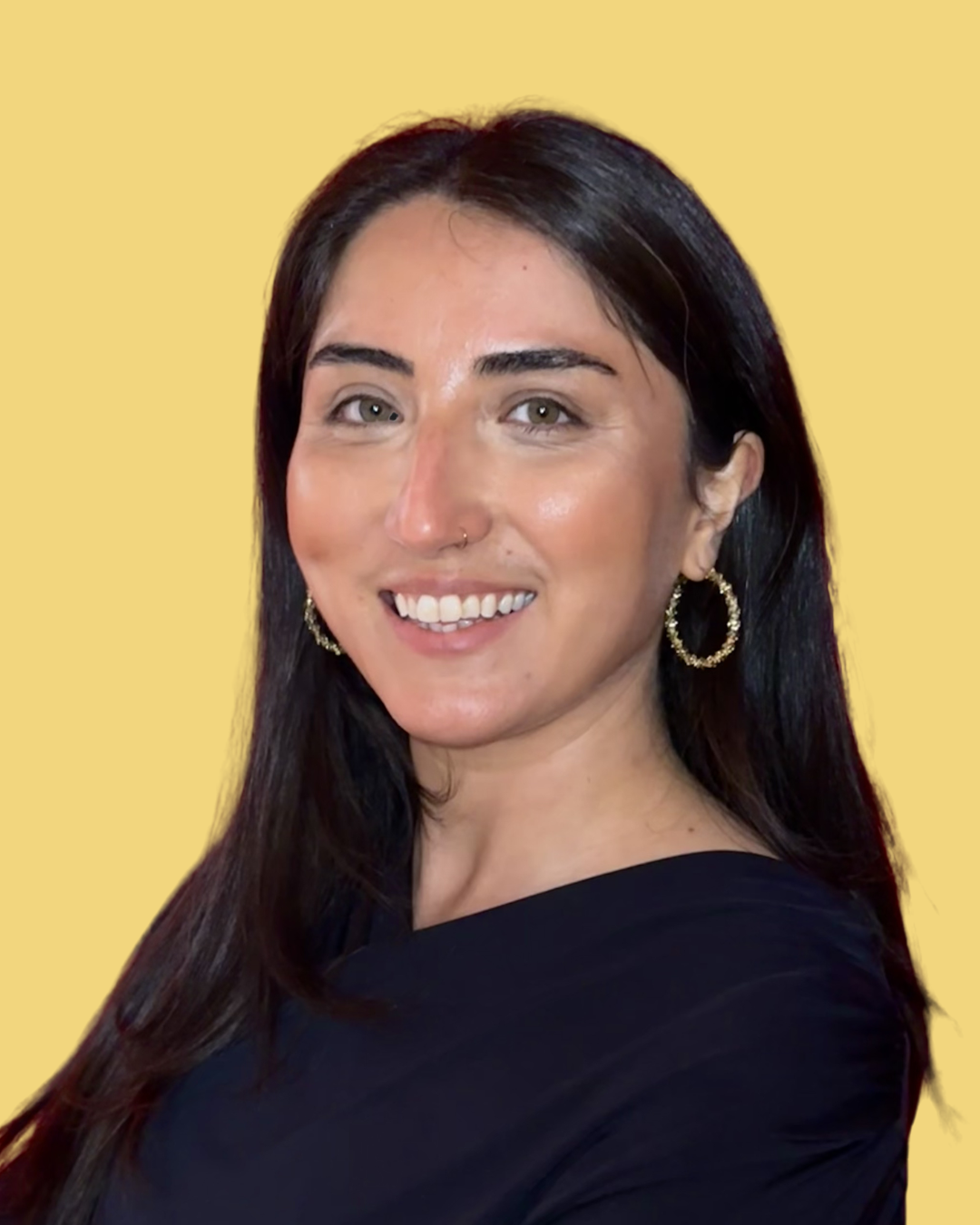 Eine lachende Frau mit schwarzen Haaren, Ohrringen und schwarzem Oberteil vor hellgelbem Hintergrund.