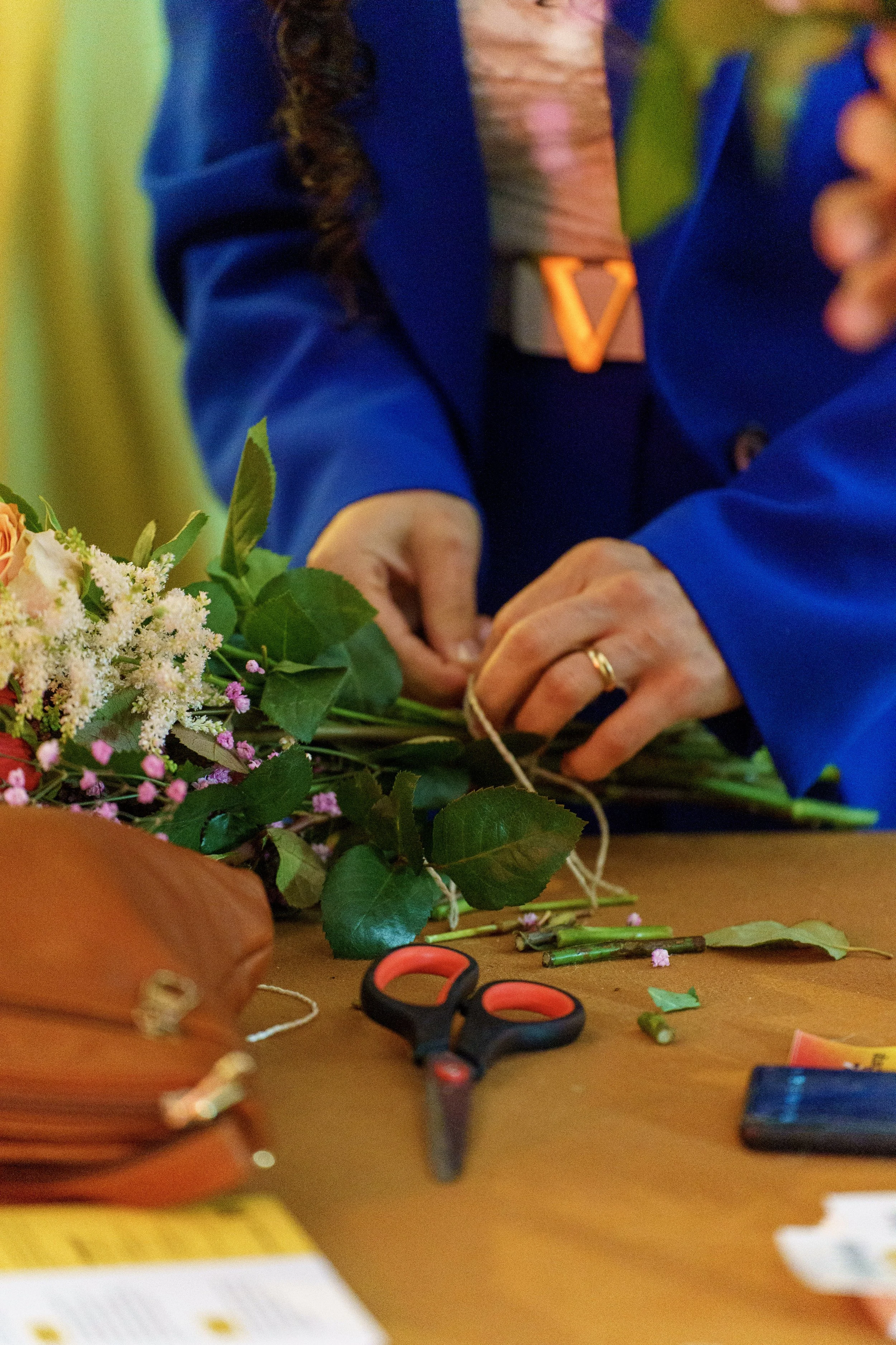 Person bindet Blumen an einem Tisch. Es liegen eine Schere, Blätter und andere Blumen auf dem Tisch.