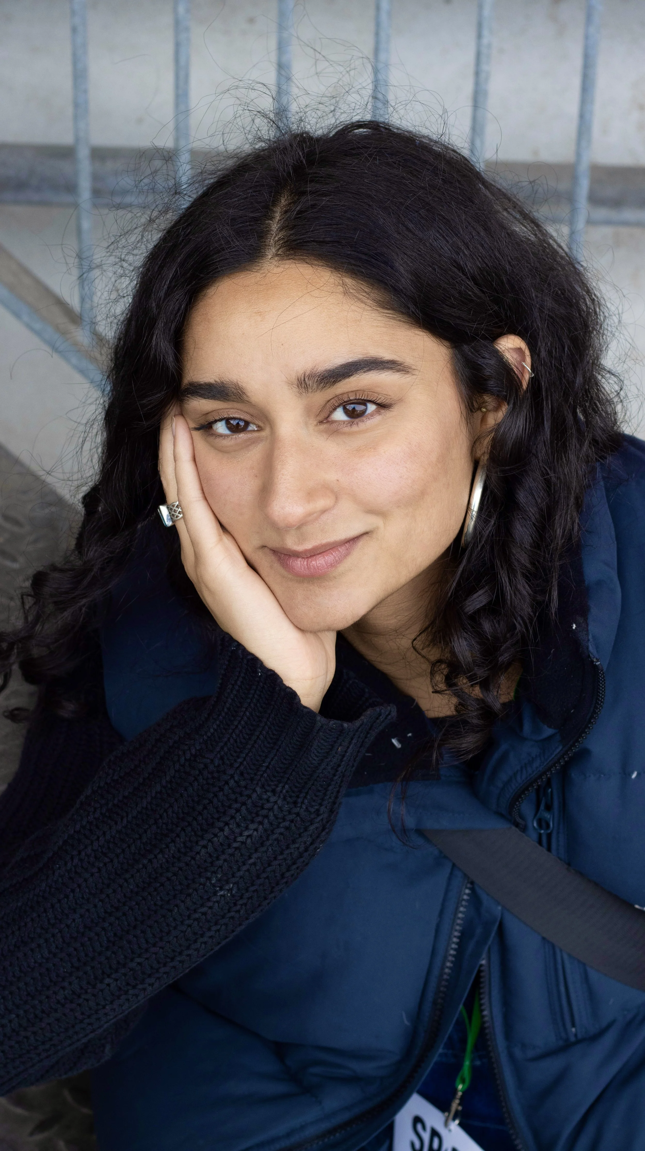 Nahaufnahme einer jungen Frau mit braunen Augen, schwarzen Locken, einem Lächeln und einer Hand, die ihr Gesicht stützt, sitzend vor einer Metallabsperrung.