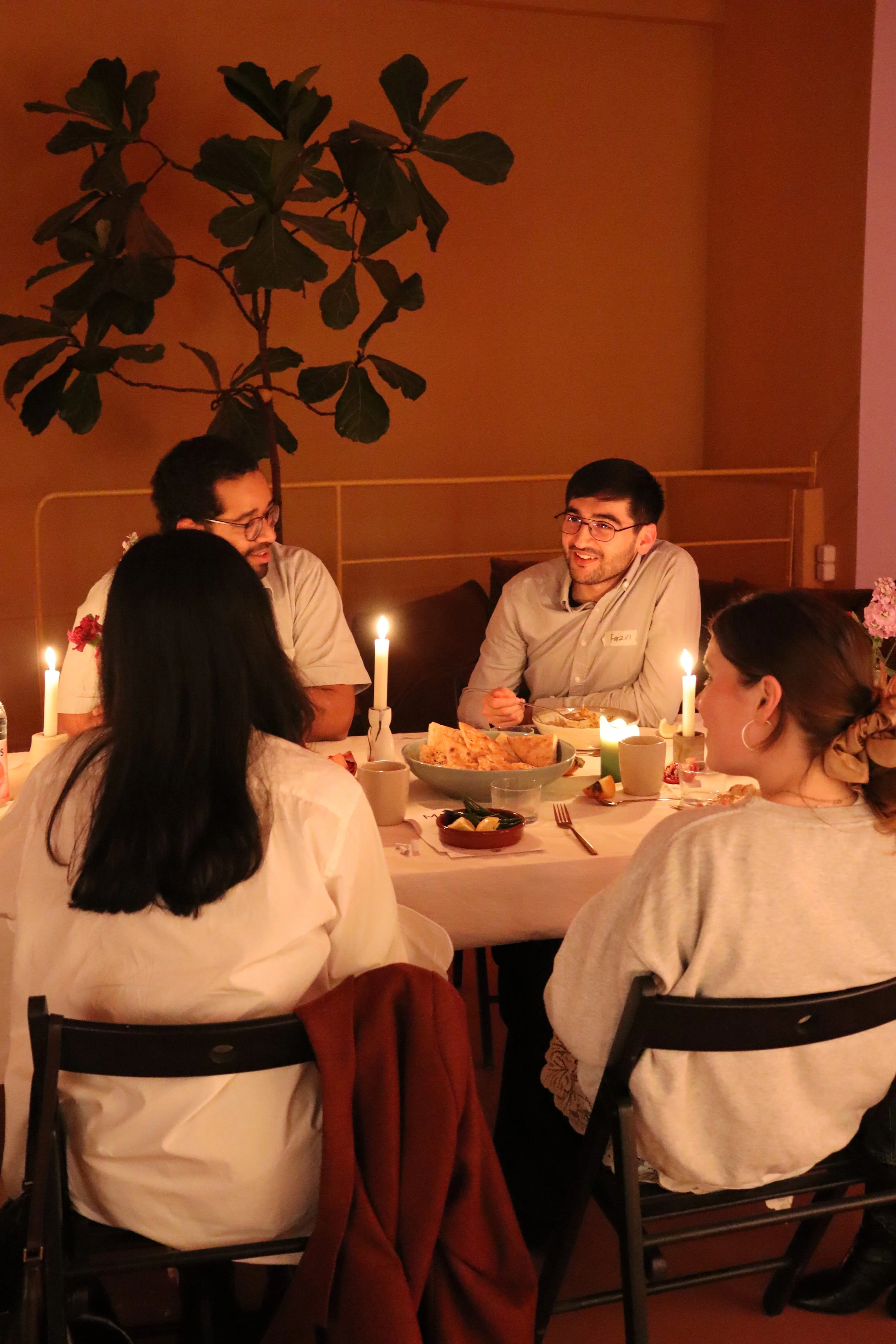 Menschen sitzen an einem festlich gedeckten Tisch bei Kerzenlicht, Essen und Getränken, im Hintergrund eine große Zimmerpflanze.