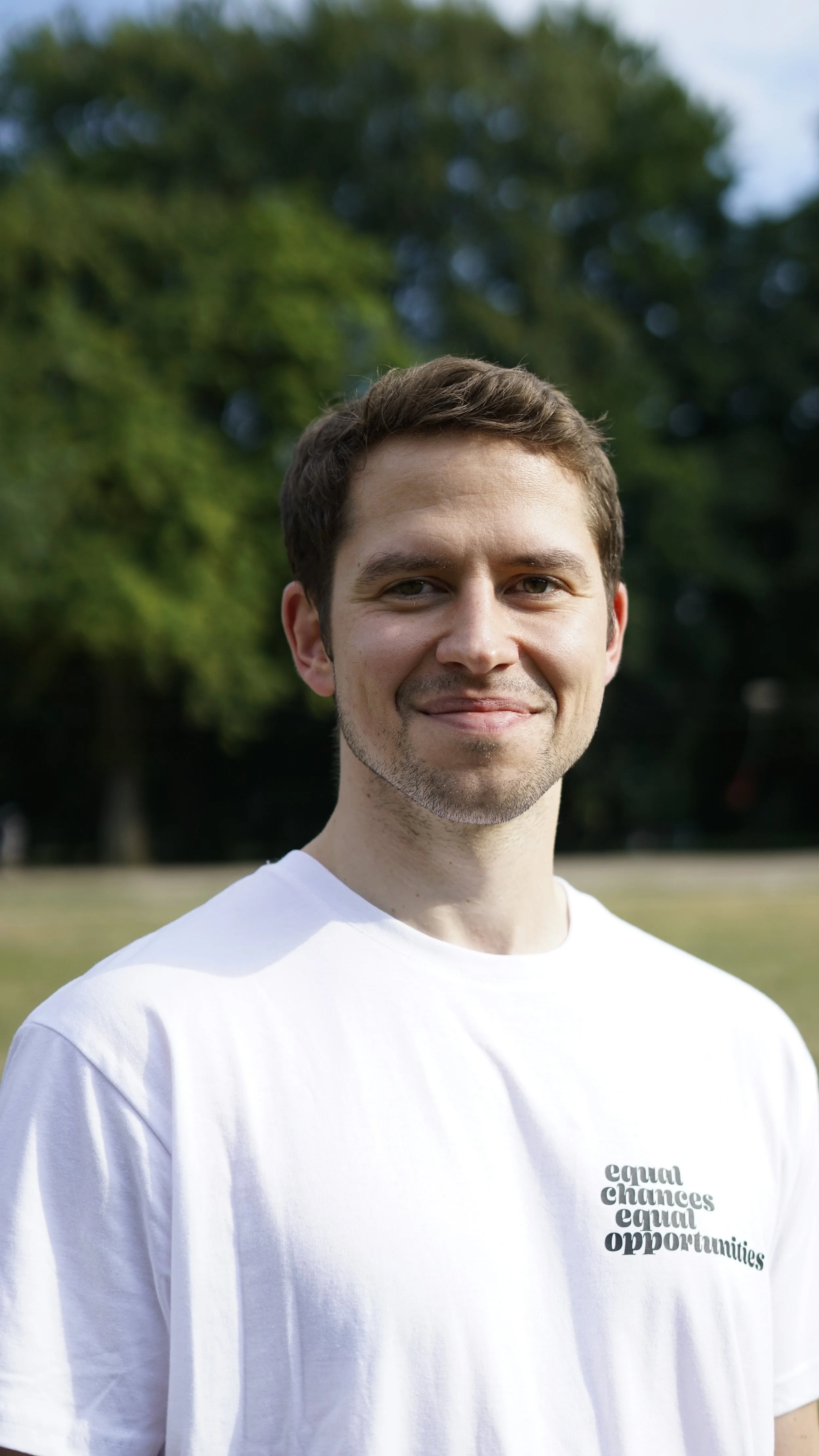 Nahaufnahme eines lachenden Mannes in einem weißen T-Shirt mit der Aufschrift „equal chances equal opportunities“ im Park, hinter ihm grüne Bäume und offener Himmel