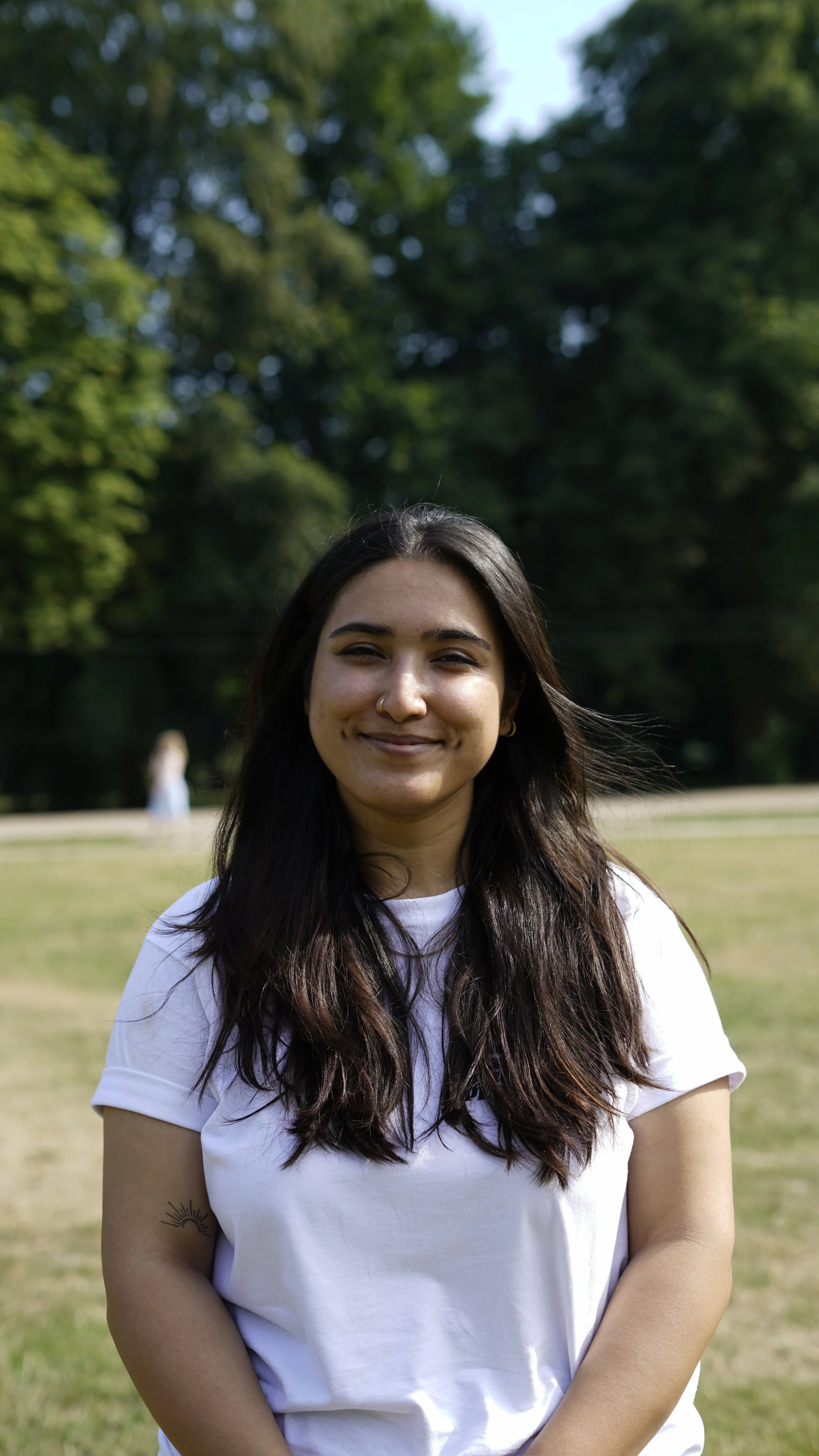 Junge Frau mit dunklen langen Haaren, weißen T-Shirt, im Park mit Bäume im Hintergrund, lächelt in die Kamera.