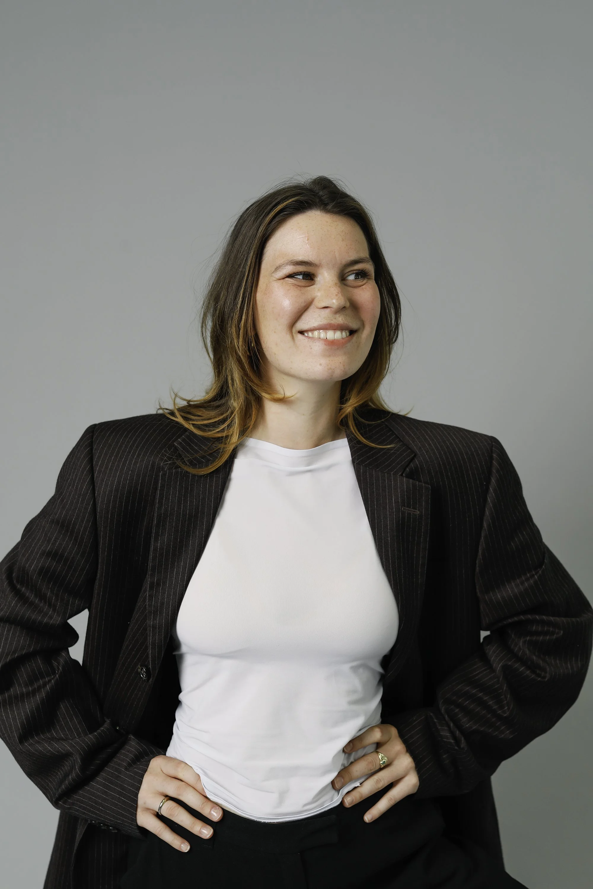 Junge Frau in Businesskleidung, lächelnd und mit Händen in die Hüften, vor neutralem grauem Hintergrund.