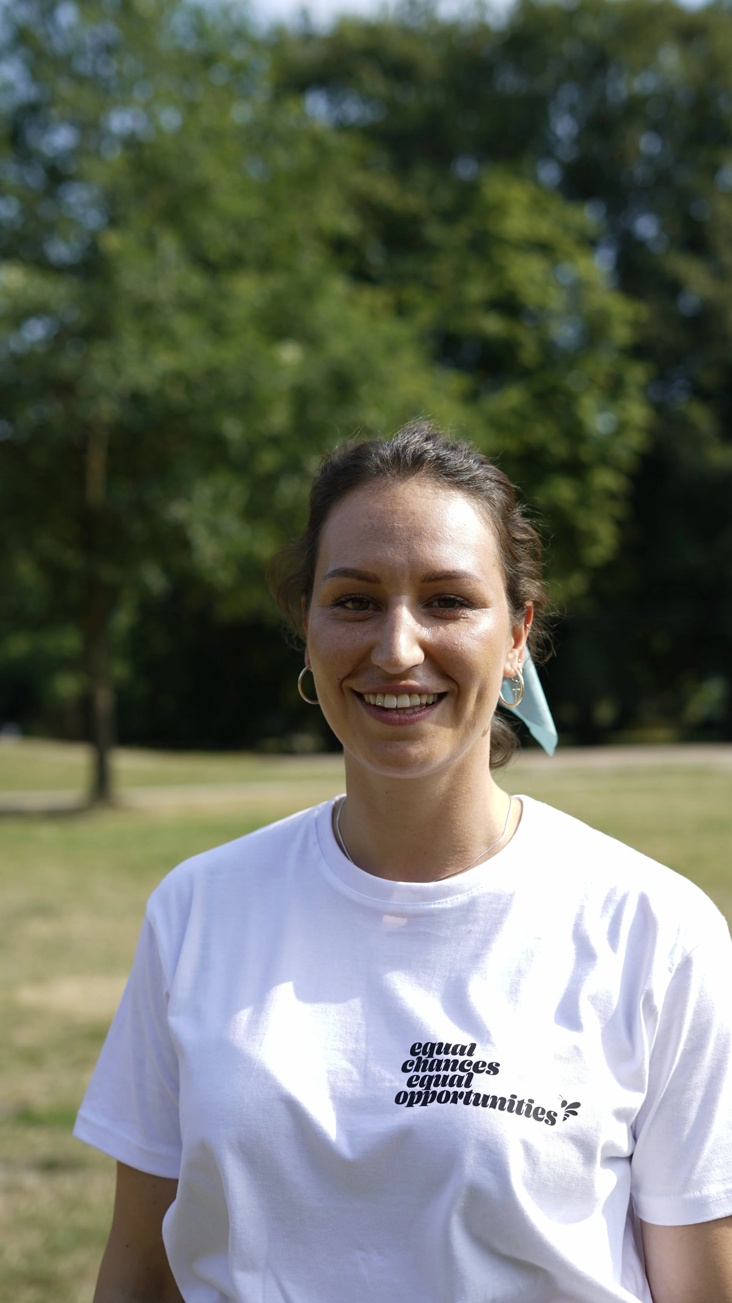 Eine lächelnde junge Frau steht im Freien in einem Park, trägt ein weißes T-Shirt mit schwarzen Texten.