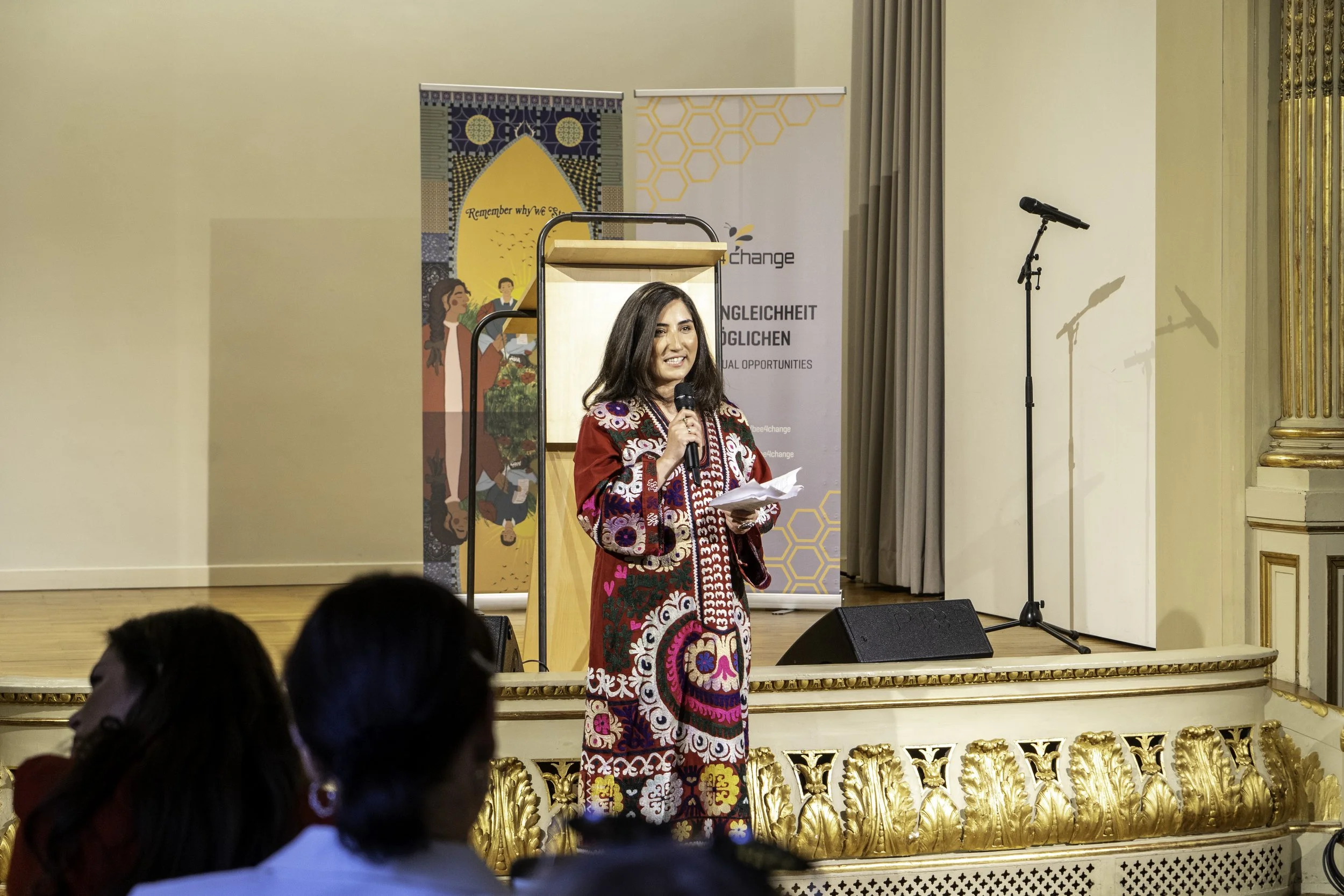 Eine Frau steht auf einer Bühne beim Reden, hält ein Mikrofon und Notizen in der Hand. Im Hintergrund sind Banner mit textuellen und grafischen Elementen sichtbar, und es gibt ein Mikrofon auf einem Ständer sowie eine goldverzierte Kulisse.