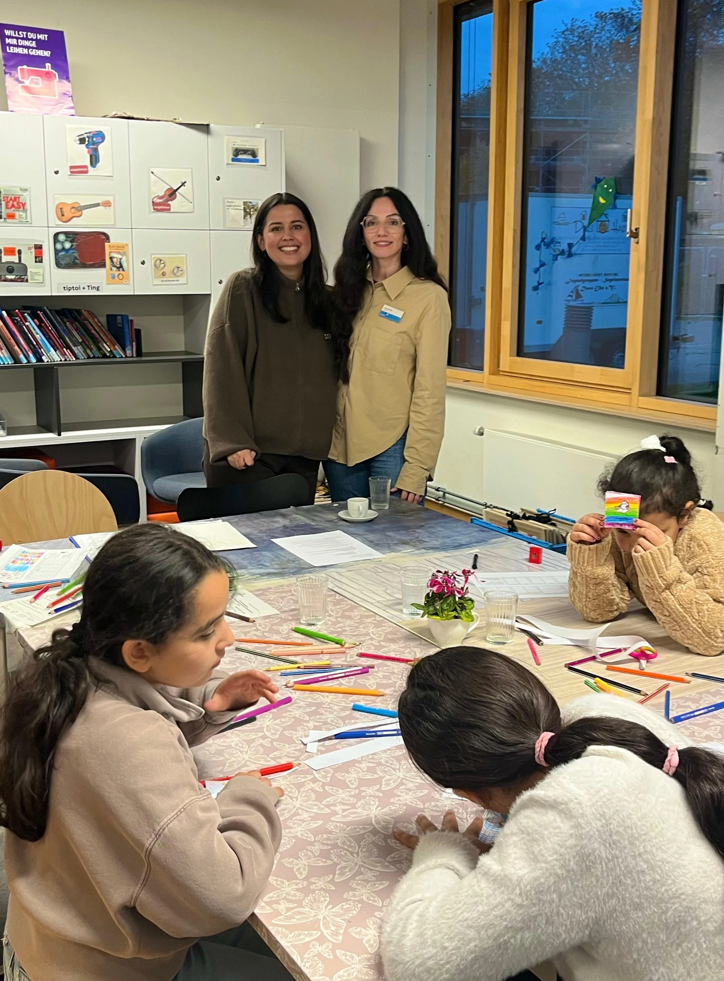 Zwei Frauen stehen hinter einem Tisch mit mehreren Kindern, die bunte Stifte und Papier haben. Das Bild scheint in einem Klassenraum oder Gemeinschaftsraum zu sein.