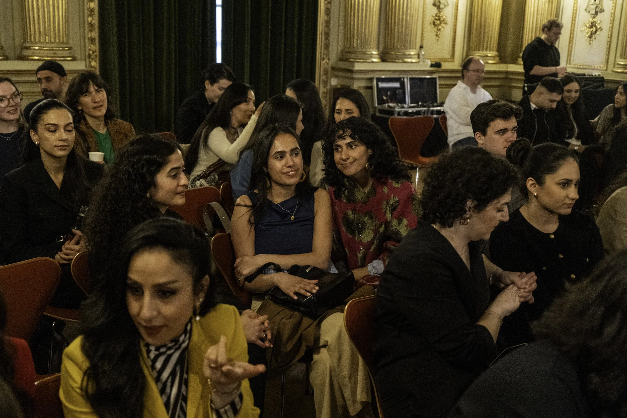 Eine Gruppe von Menschen sitzt in einem eleganten, goldverzierten Raum bei einer Veranstaltung, einige unterhalten sich, andere schauen aufmerksam.