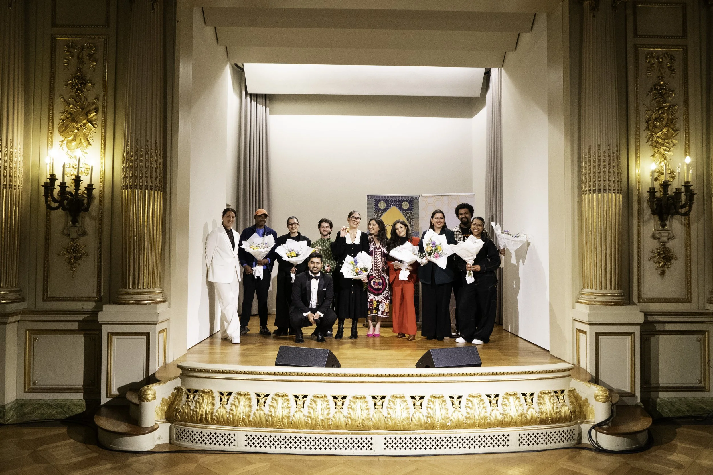 Gruppe von Menschen auf einer Bühne, einige tragen Blumen, in einem eleganten Saal mit goldverzierten Säulen und Wanddekorationen.