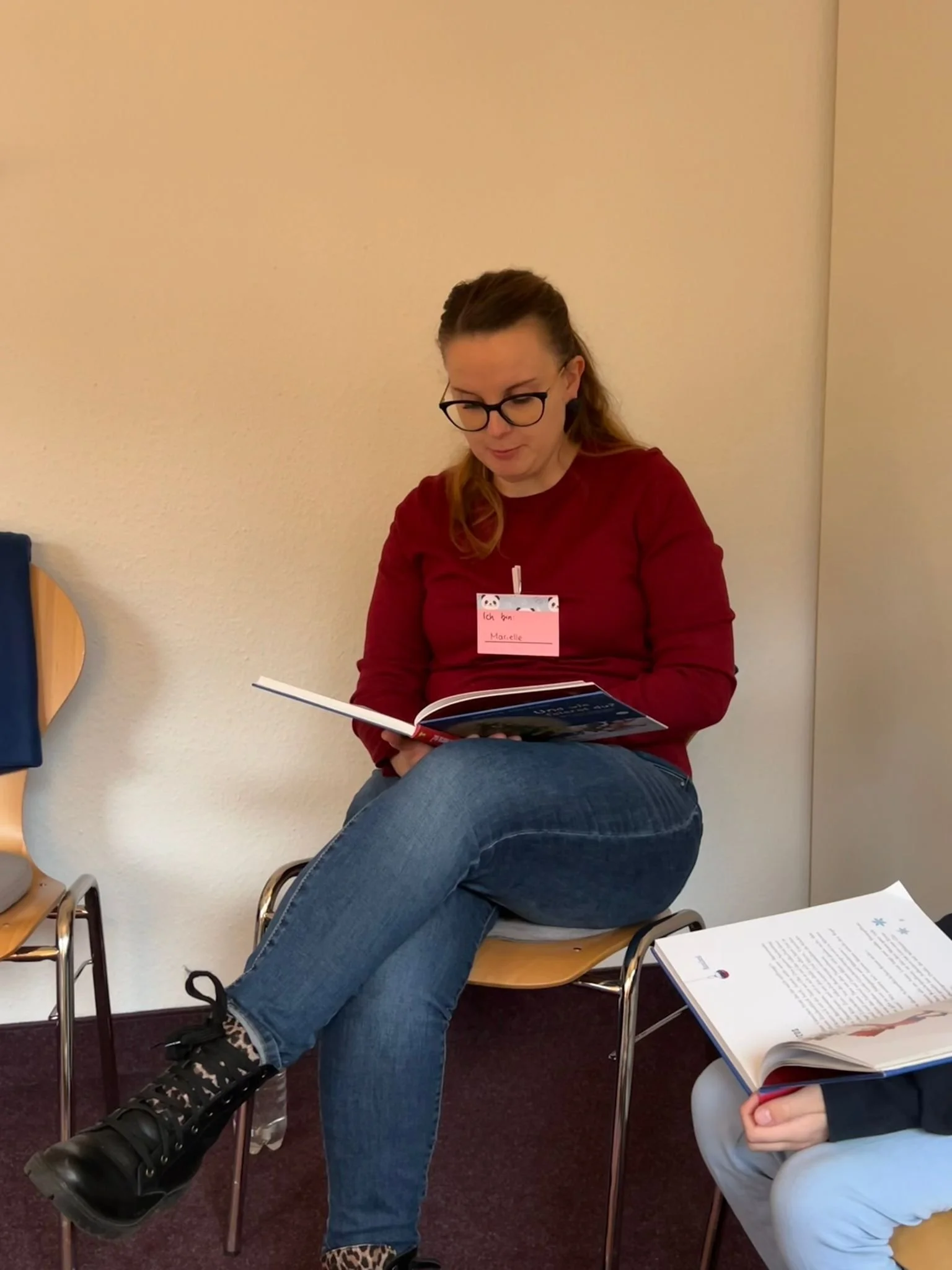 Eine Frau sitzt auf einem Stuhl und liest ein Buch, trägt eine rote Bluse und Jeans, mit Brille und Namensschild am Hals, im Hintergrund eine weiße Wand.