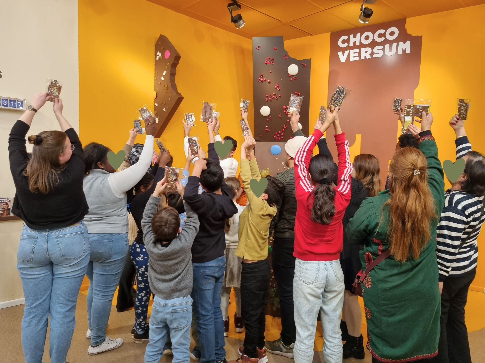 Menschenmenge beim choco versum, alle mit Schokoriegeln in der Hand, vor einer orangefarbenen Wand mit Schokoladen-Design, einige Kinder und Erwachsene, bunt gekleidet, feiern.