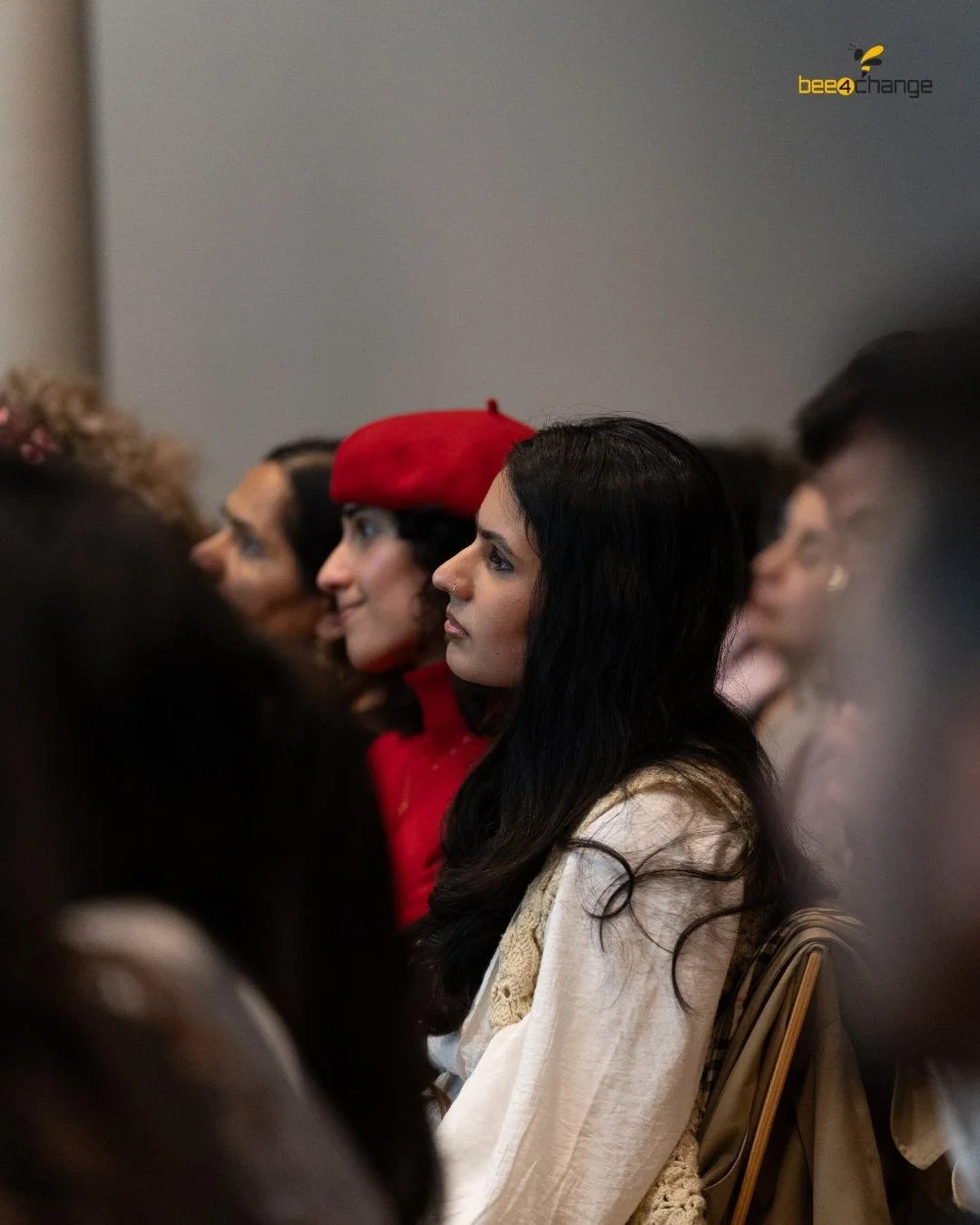 Eine Gruppe von Menschen, darunter eine Frau mit langen schwarzen Haaren in traditioneller Kleidung, sitzt vertieft bei einem Vortrag oder einer Veranstaltung, im Hintergrund ein Mann in rotem Hut und weiterer Personen.