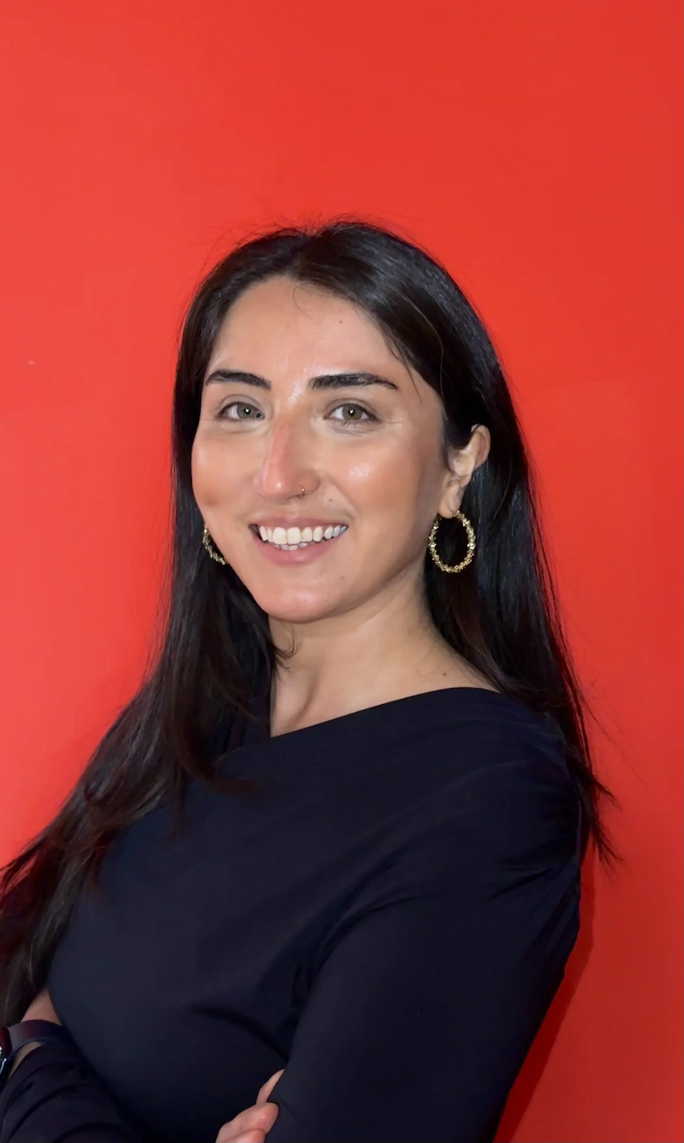 Eine lächelnde Frau mit schwarzen Haaren, Gold-Ohrringen und dunklem Top vor einem roten Hintergrund.