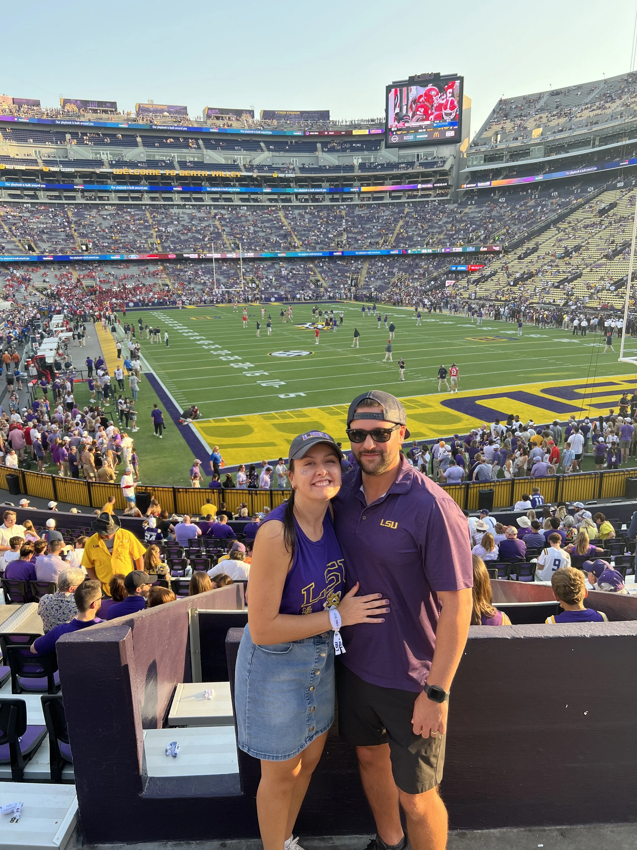 A young woman and an older man stand together at a football stadium, smiling for the photo. Both are wearing LSU merchandise, with the woman in a purple LSU shirt and the man in a purple LSU polo. The stadium is filled with fans, and the field is visible in the background.