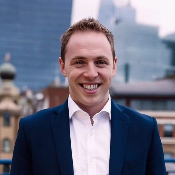 A smiling man in a navy blue suit and white shirt, with city buildings in the background.