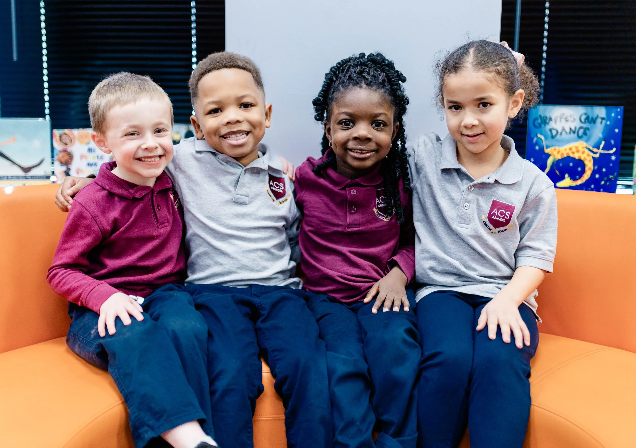 Four diverse children sit closely together on an orange couch in a classroom, smiling at the camera, wearing school uniforms with a badge reading 'ACS'.