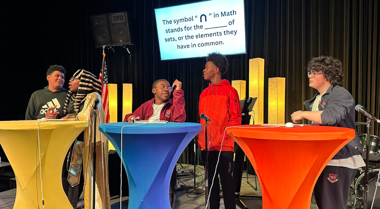 Children on stage participating in a quiz or discussion, with colorful tables, microphones, and an American flag. A screen displays a question about the symbol '∩' in math.