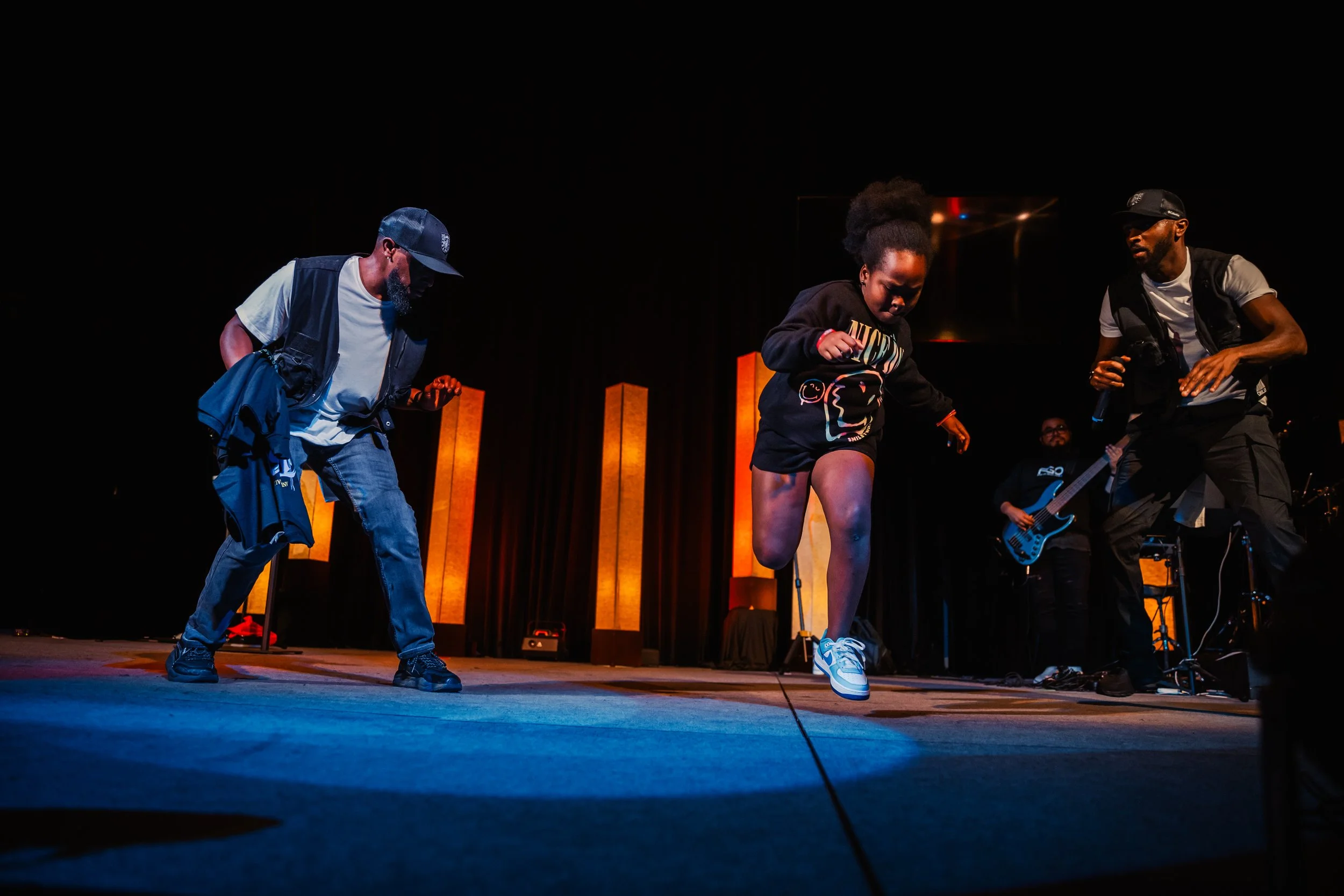 People dancing on stage during a live performance, with a musician playing guitar in the background and illuminated orange vertical light panels.