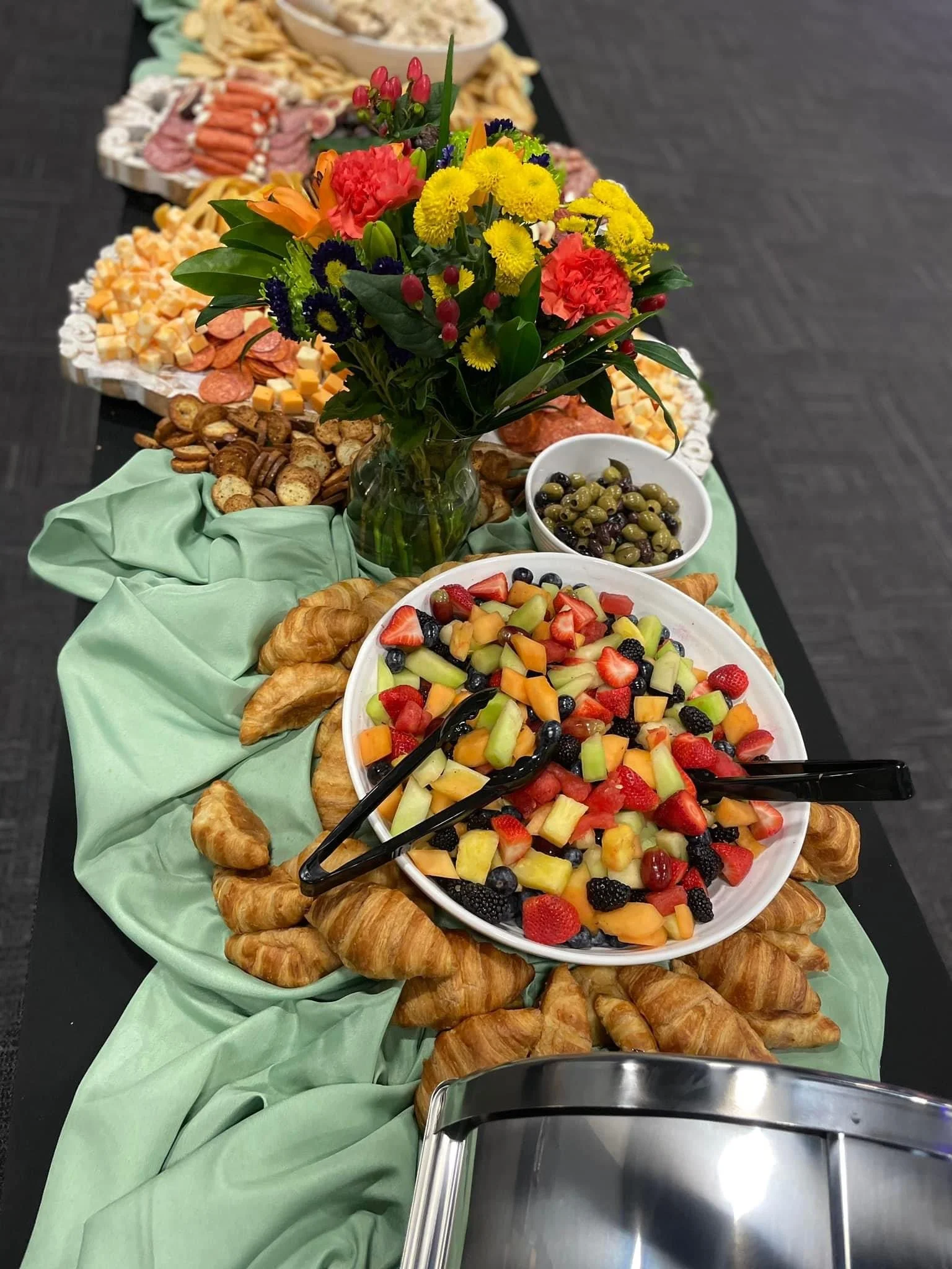 A festive table with a green tablecloth featuring an assortment of fruits, pastries, cheese, nuts, olives, and a colorful flower arrangement, set up for a buffet.
