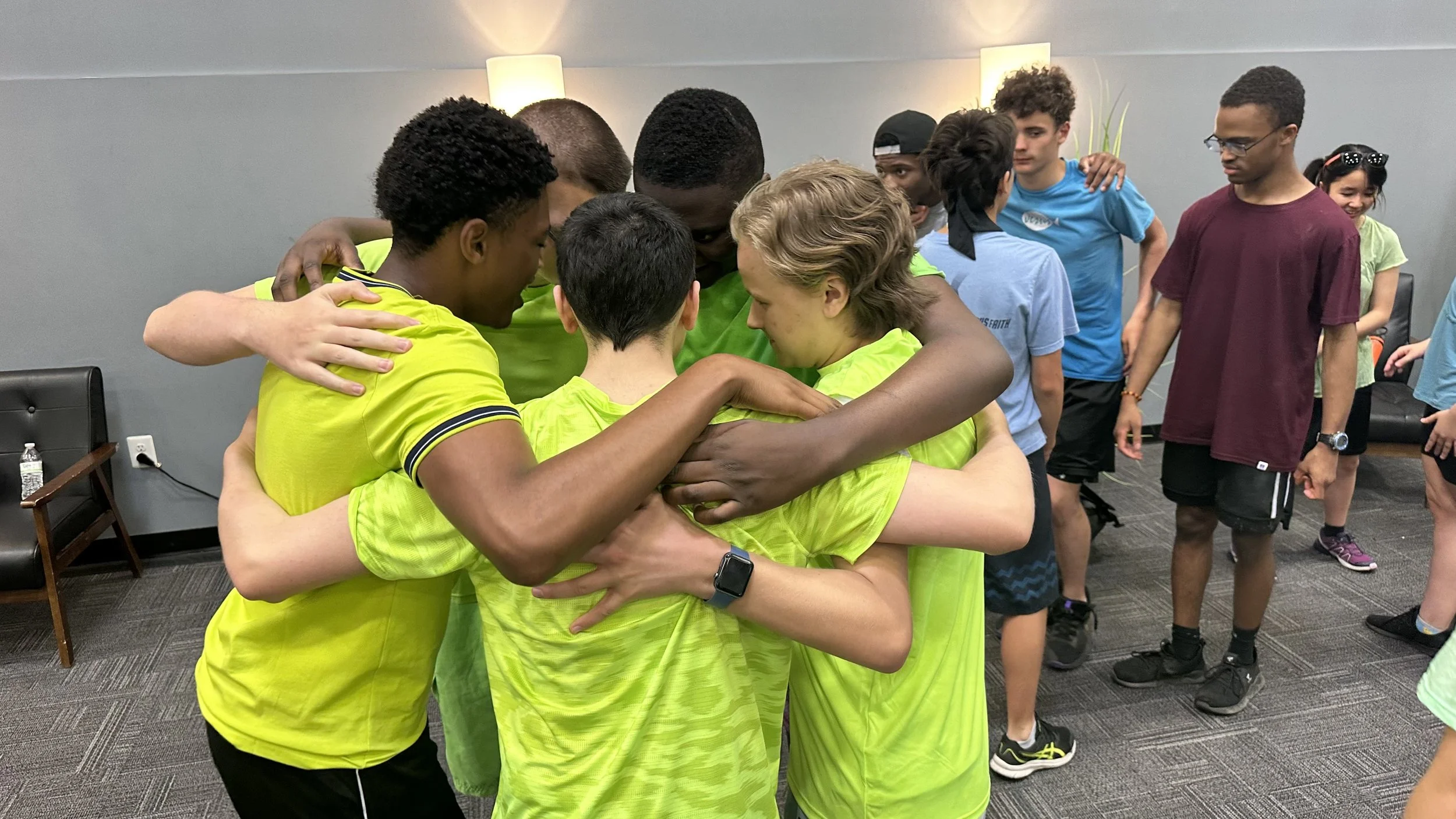 A group of young people in sports wear hugging in a circle in a room with gray walls and black chairs.