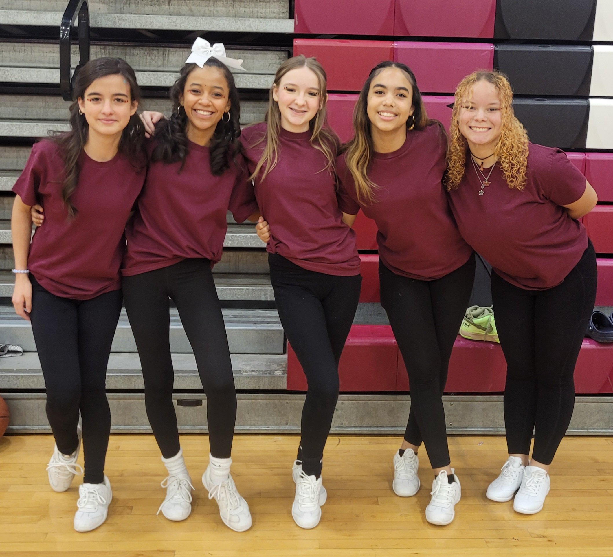 Five young women standing together in a gym, posing with arms around each other, wearing maroon t-shirts, black pants, and white sneakers.