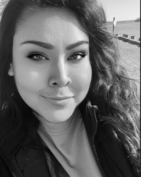 A close-up black and white photo of a woman with long, wavy hair, smiling slightly, with a blurred outdoor background.