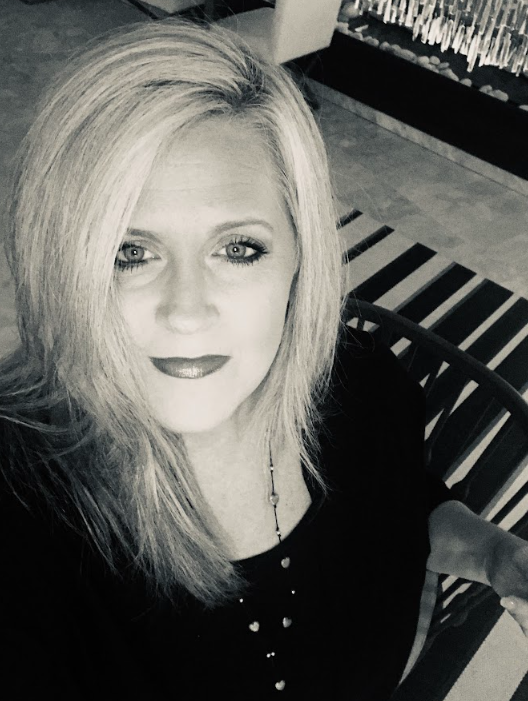 A woman with blonde hair taking a selfie, sitting on a bench in a well-lit indoor space.