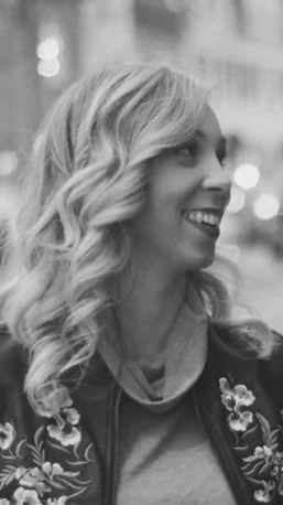 A woman with wavy blonde hair smiling, wearing a floral jacket and a gray hoodie, outdoors in a city setting.