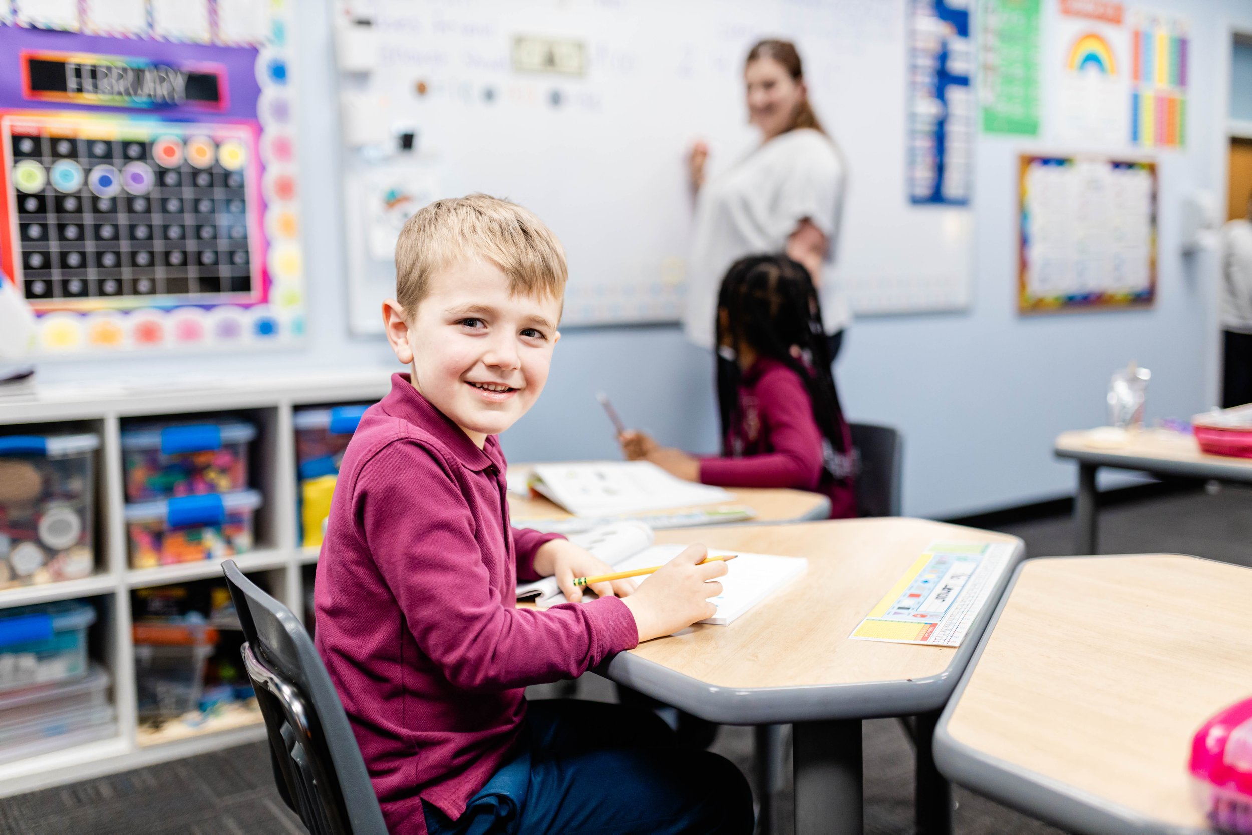 A young boy sitting at a classroom desk, smiling at the camera, with a teacher and another student in the background.